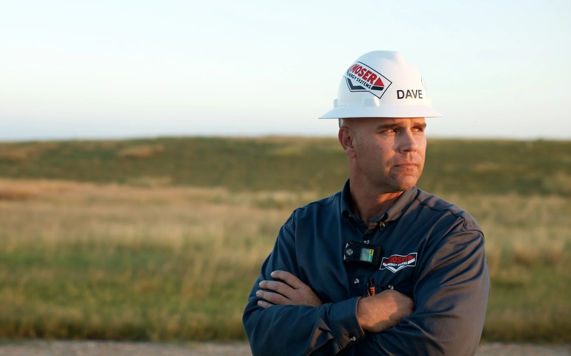 Man in a navy uniform with a hard hat and safety equipment, standing outdoors with arms crossed, in a field during sunset.