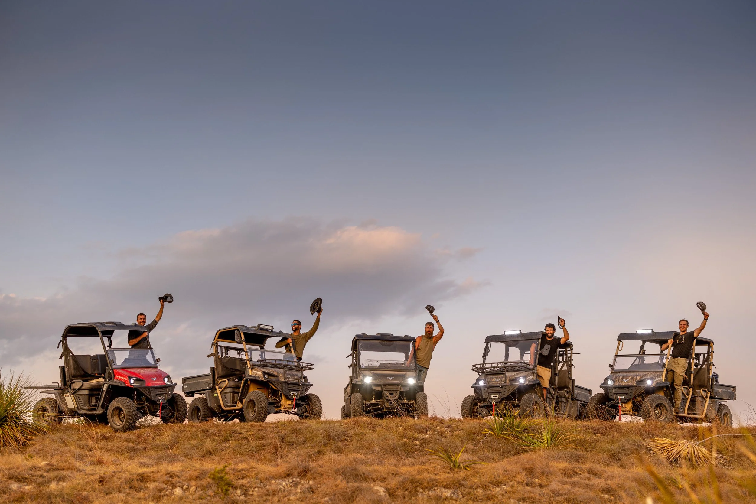 Six men standing in off-road utility vehicles on a grassy hilltop during sunset, waving hats in the air.