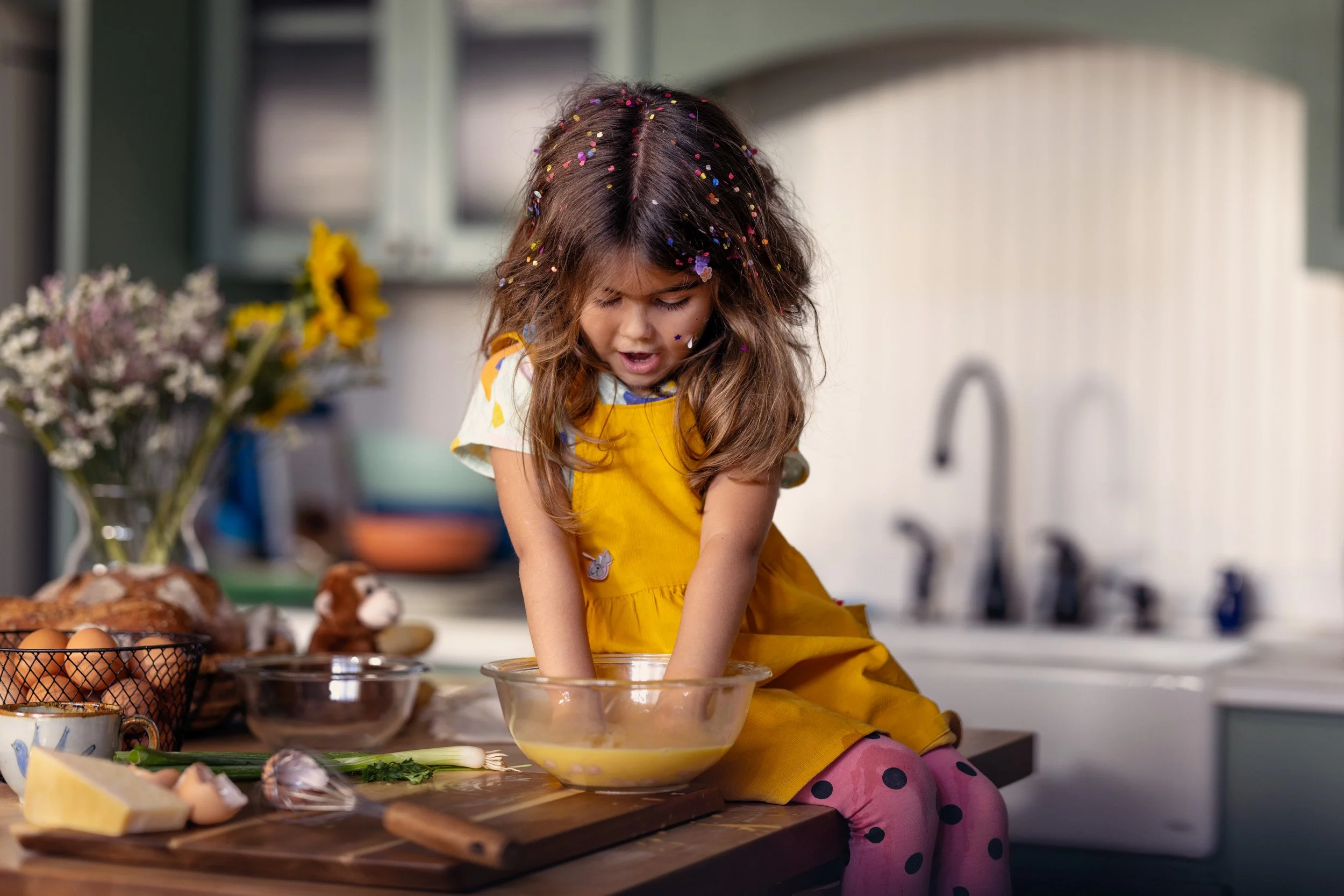A young girl with brown hair covered in colorful confetti, wearing a yellow apron and pink polka-dotted leggings, is mixing ingredients in a glass bowl on a kitchen countertop. There are eggs, a loaves of bread, and other cooking ingredients on the c
