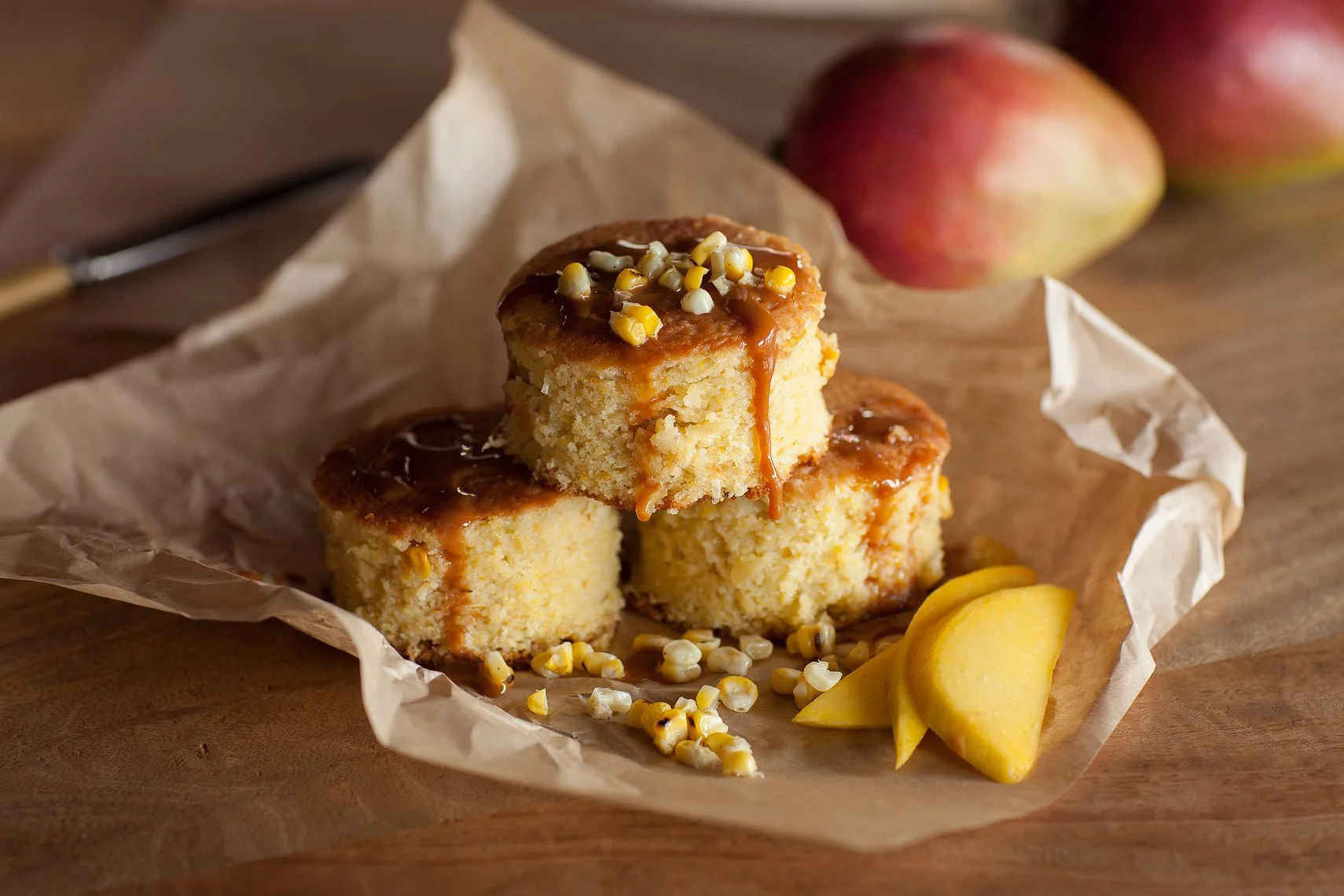 Three slices of cake with caramel sauce and corn topping on parchment paper, with sliced mango and whole apples in background on a wooden surface.