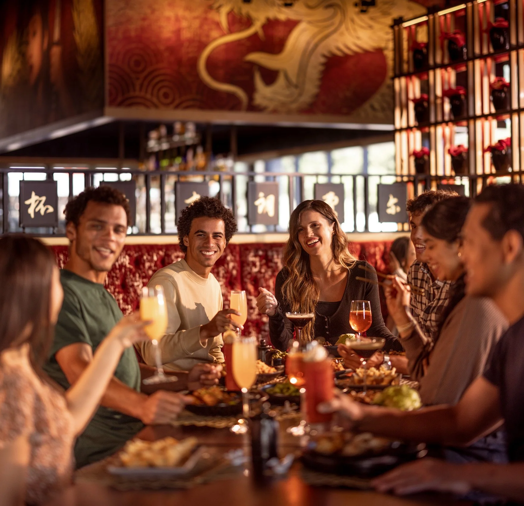 A group of people dining together at a restaurant. They are smiling and holding drinks, with food and beverages on the table. The background features warm lighting, a decorative wall with a dragon artwork, and shelves with rose decorations.
