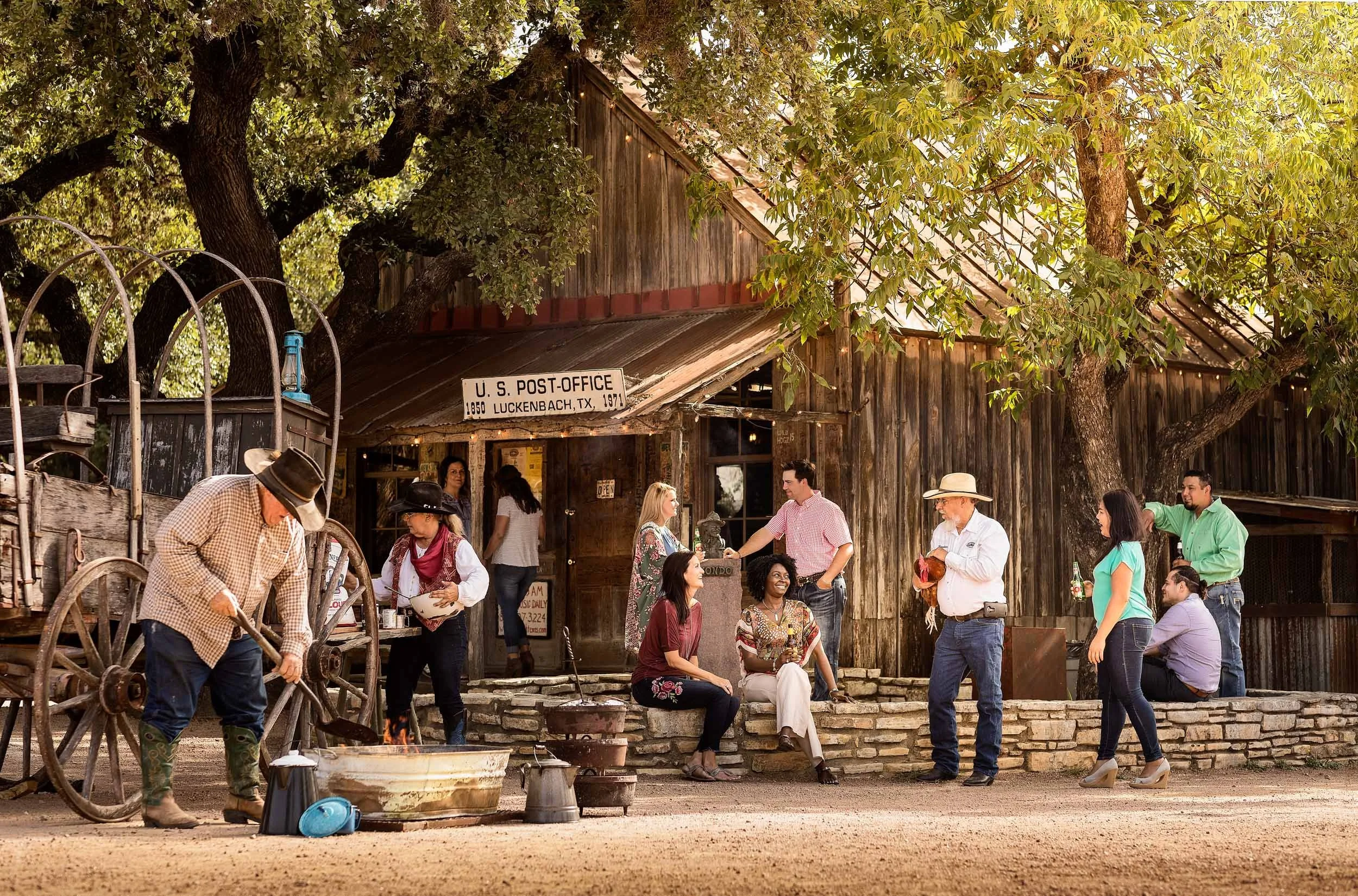 People gathered outside a rustic wooden building with a sign saying U.S. Post Office, Luckenbach, TX, 1971. Some are sitting, others standing, with a man tending a fire in the foreground, and a large tree with green leaves overhead.