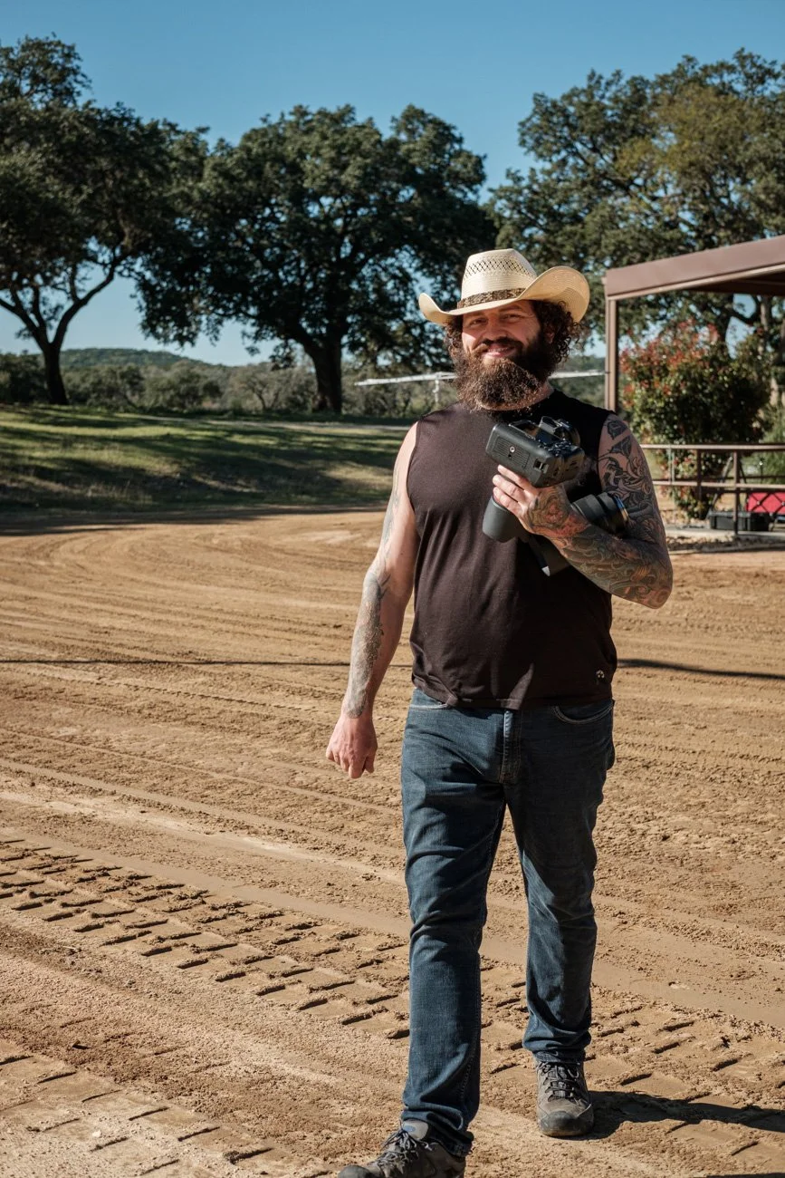 A man with a beard and tattoos wearing a cowboy hat, black sleeveless shirt, and jeans, holding a camera, standing on a dirt path at a park or outdoor area with trees and a pavilion in the background.