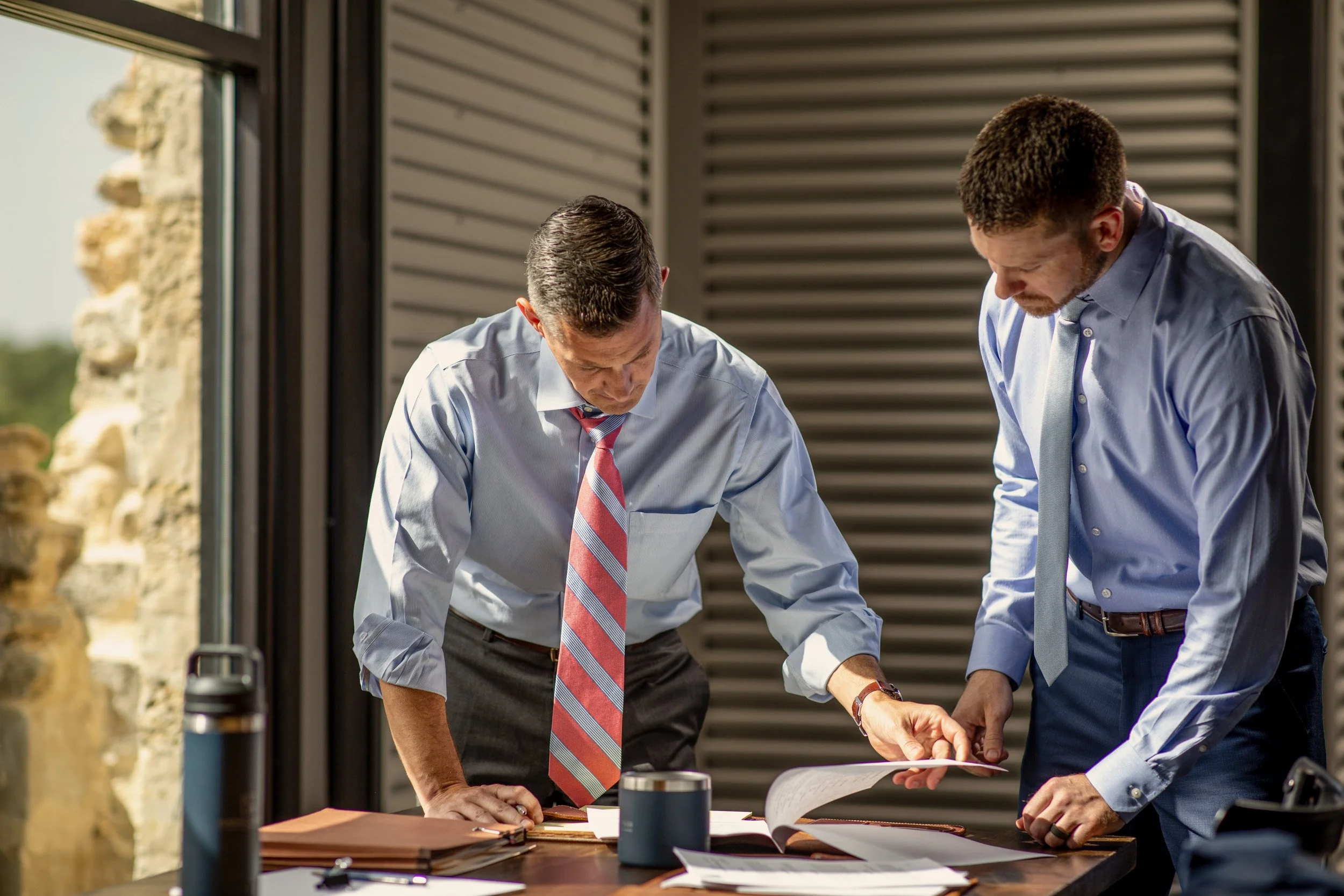 Two men in business attire examining documents at a table outdoors, with a modern building and stone wall in the background.