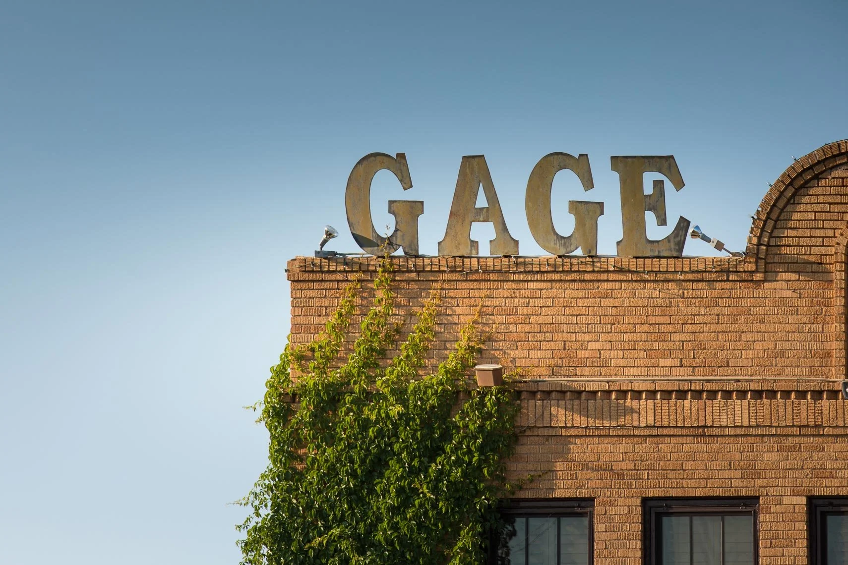 Close-up of a brick building with a large metal sign spelling 'GAGE' on the rooftop, surrounded by green vines against a clear blue sky.