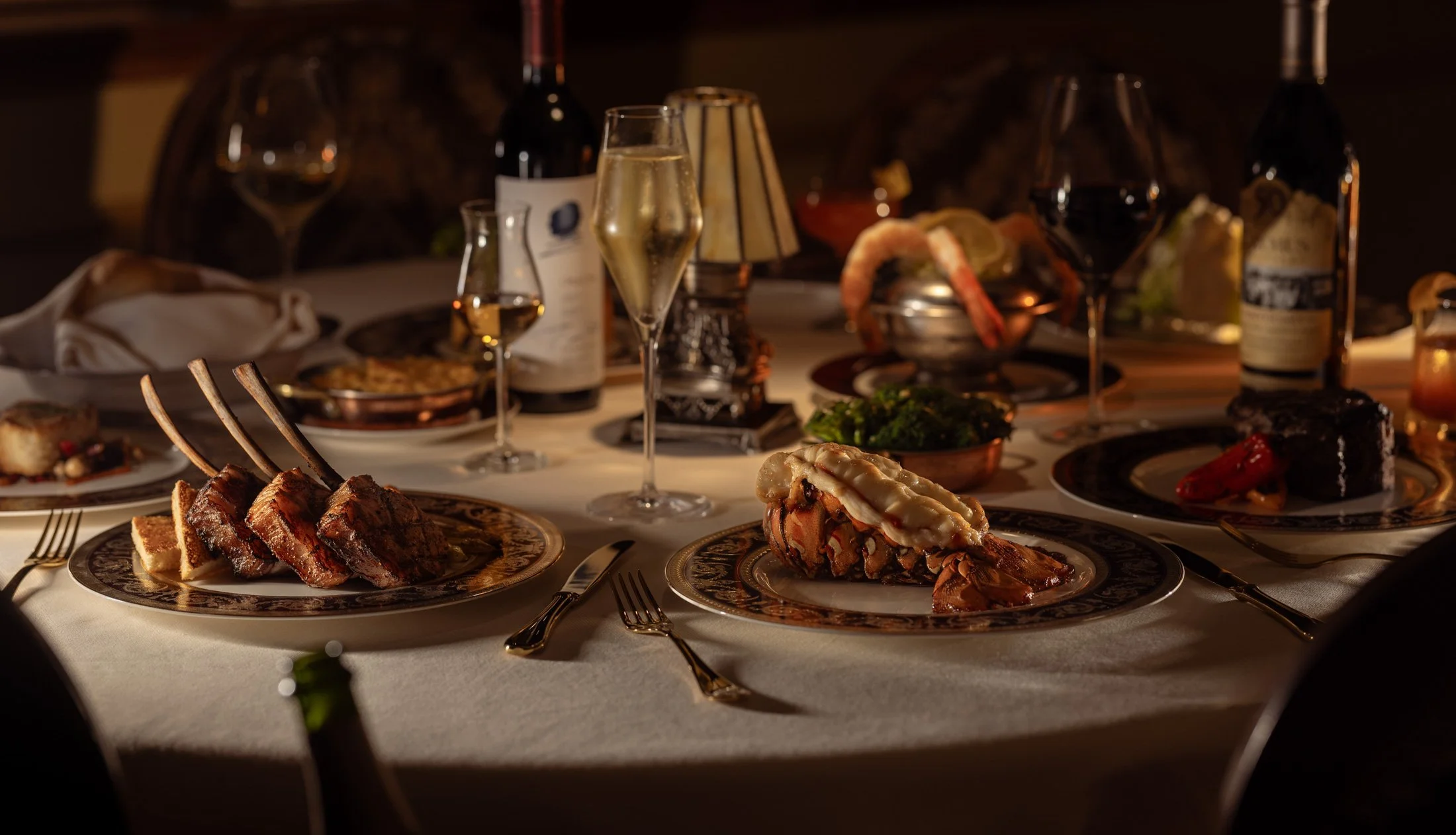 A family dinner table set with various dishes including steak, seafood, desserts, and glasses of wine, with warm lighting.