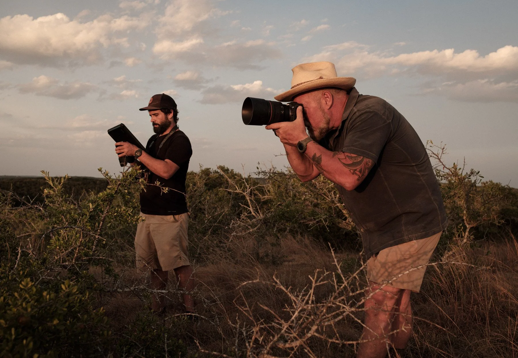 Two men in a natural outdoor setting, one taking photos with a camera and the other looking at a device, both surrounded by bushes and low vegetation under a partly cloudy sky.