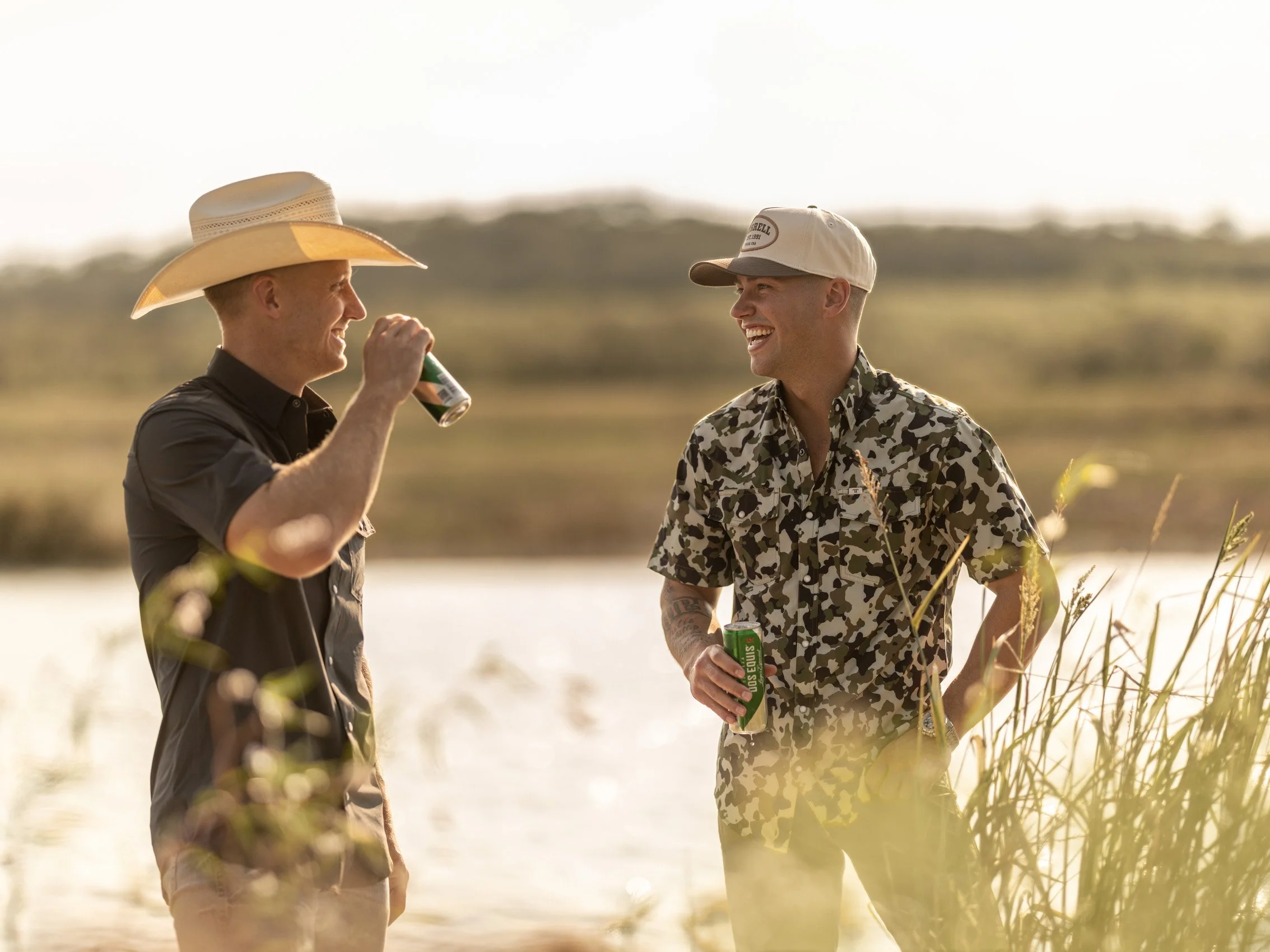 Two young men are standing outdoors near a body of water, smiling and holding drinks. The man on the left is wearing a wide-brimmed straw hat and a dark shirt, while the man on the right is wearing a white baseball cap and a camouflage shirt. The sce