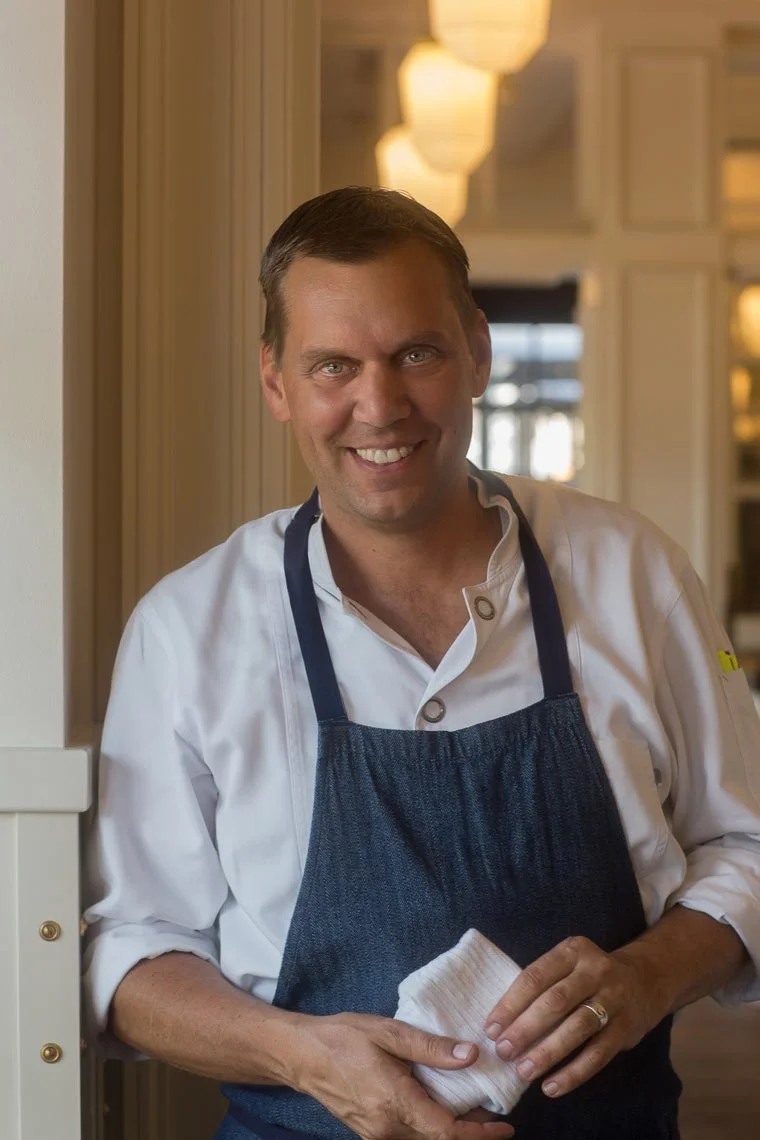 A smiling male chef in a white chef coat and dark apron holding a cloth, standing in a warmly lit restaurant kitchen.
