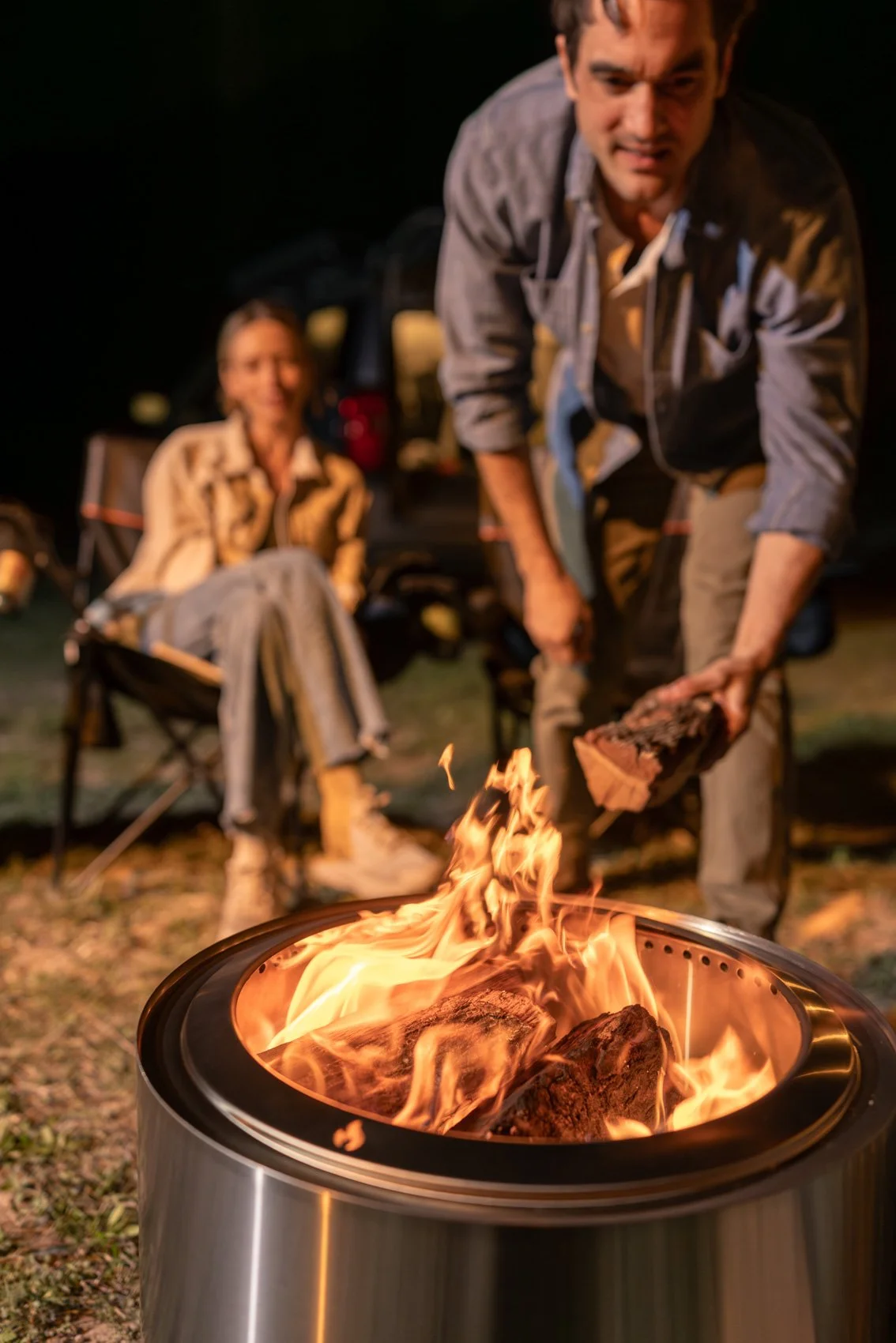 A person tending a campfire with a woman sitting in a camping chair in the background at night.