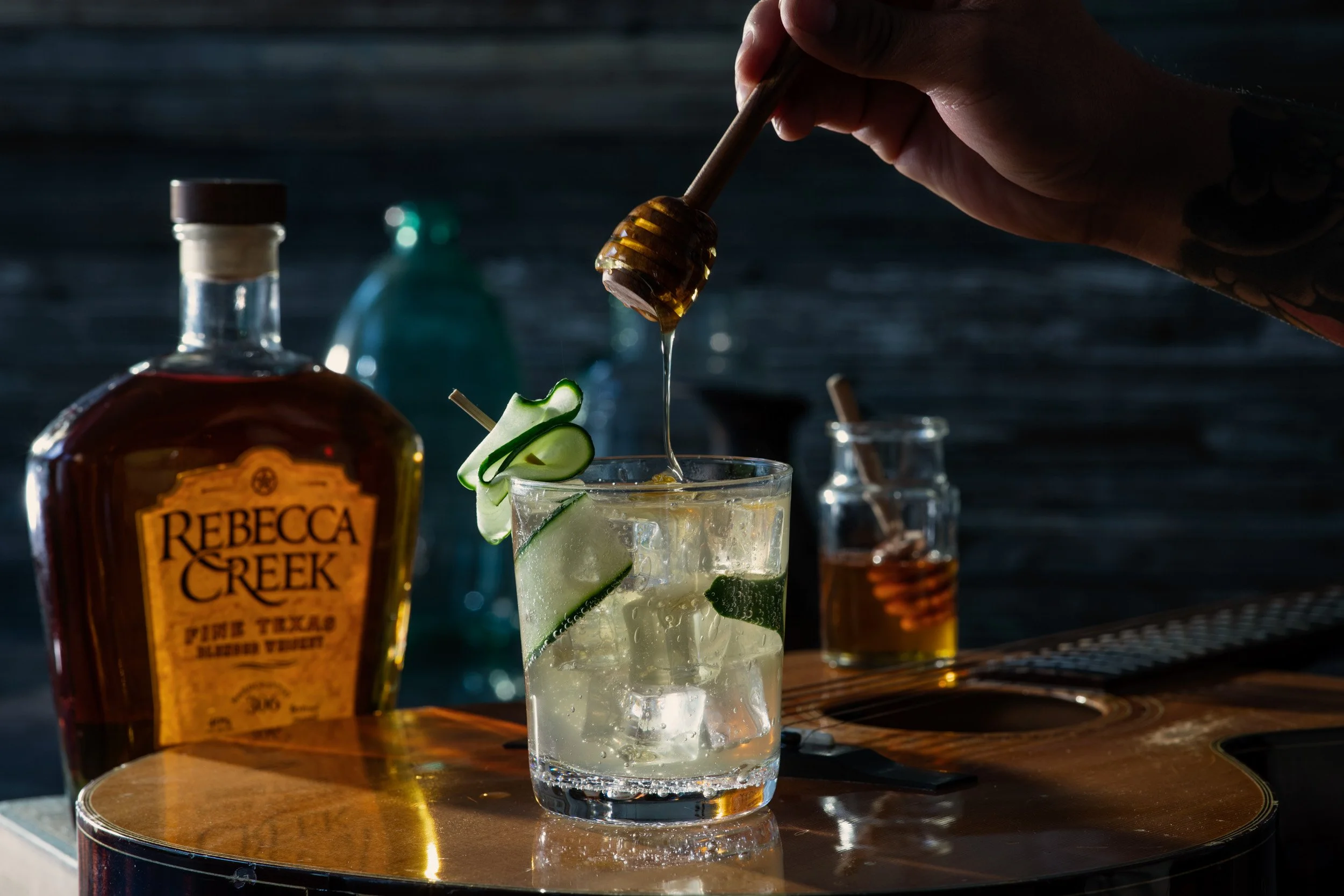 A glass of cocktail garnished with cucumber slices, honey dipper dripping honey, and bottles of liquor on a wooden table against a dark background.