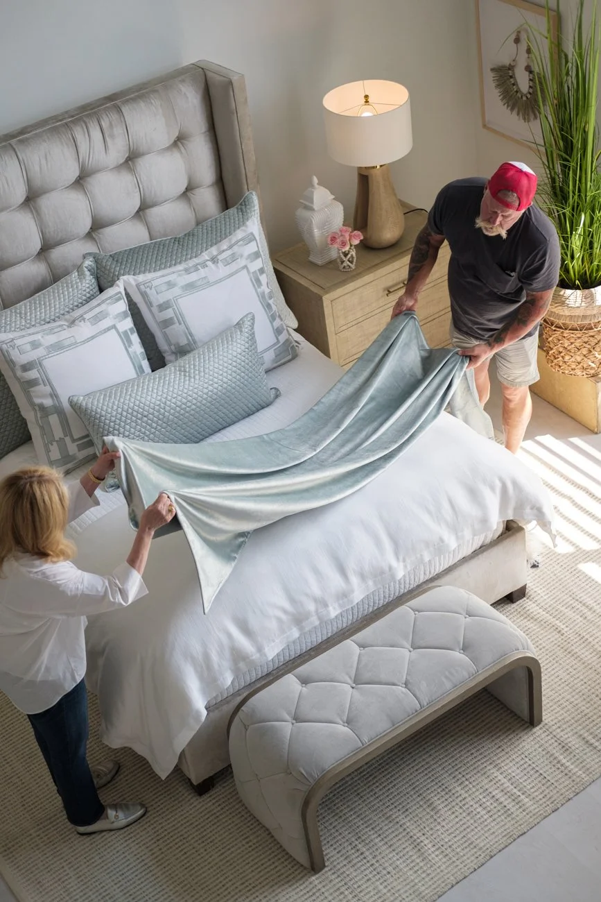 A man and woman make the bed together in a well-lit bedroom with a tufted headboard, decorative pillows, a nightstand with a lamp, vase, and flowers, a large potted plant, and artwork on the wall.