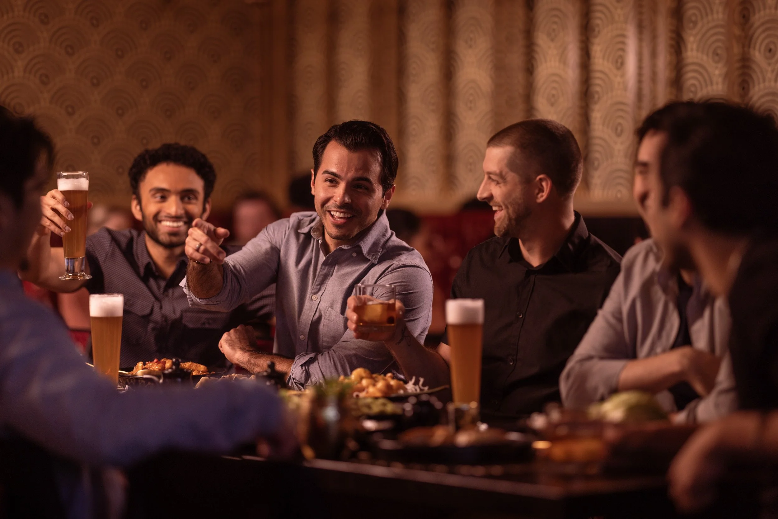 Group of friends enjoying drinks and food at a social gathering in a restaurant or bar, with a warm, cozy ambiance and patterned wallpaper.