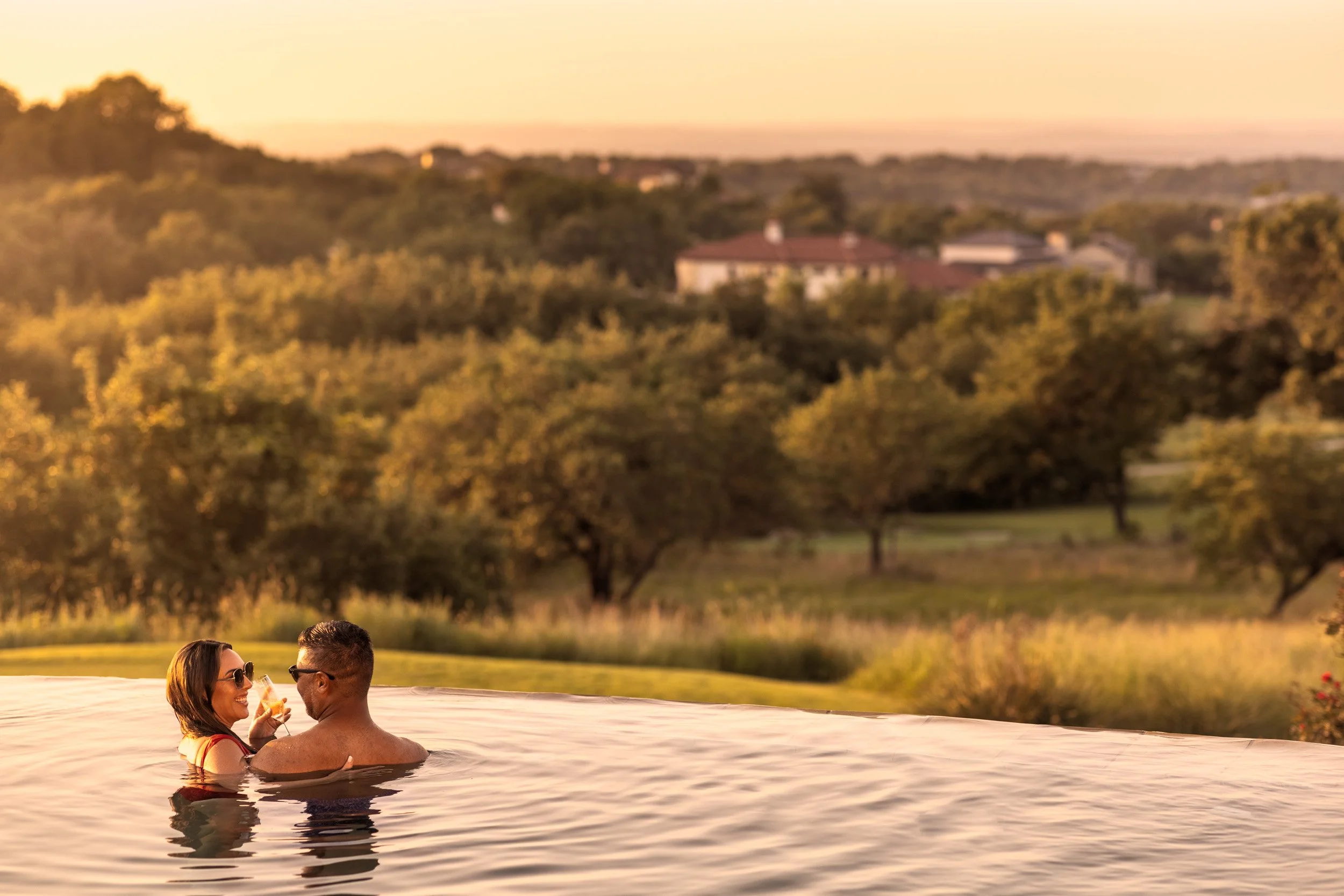 A couple is happily enjoying a swim in a pool during sunset, with a scenic view of trees and houses on rolling hills in the background.