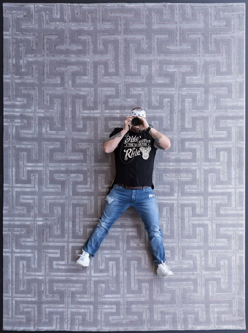 Person taking a photo of himself in a mirror with a camera, standing on a patterned grey rug.