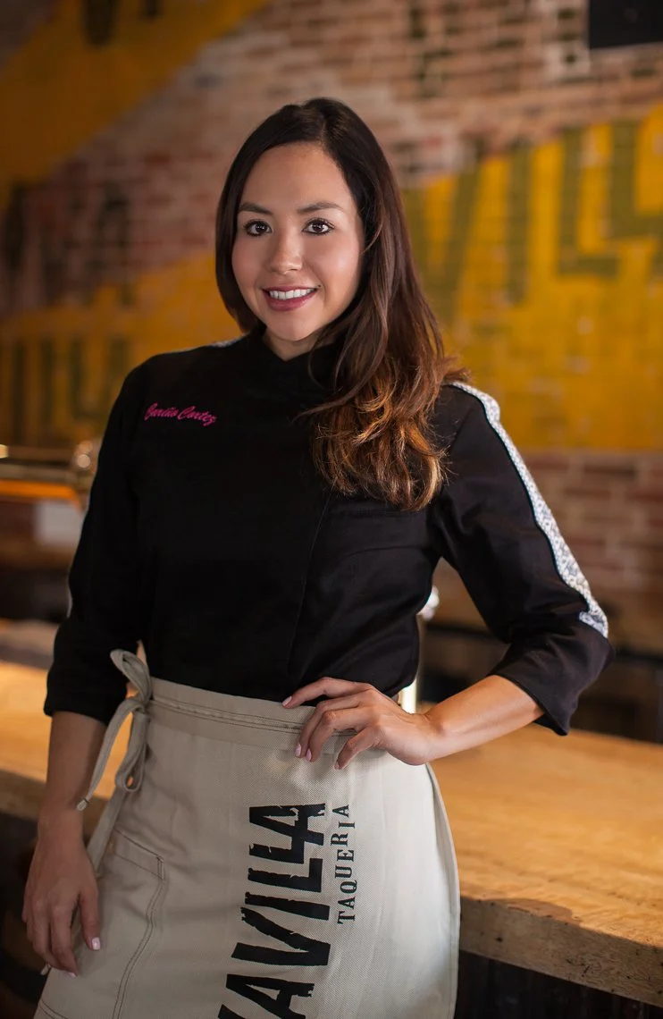 A woman with long brown hair wearing a black chef's coat with a name embroidered in pink and a tan apron with black text, standing inside a restaurant with exposed brick walls and yellow signage in the background.