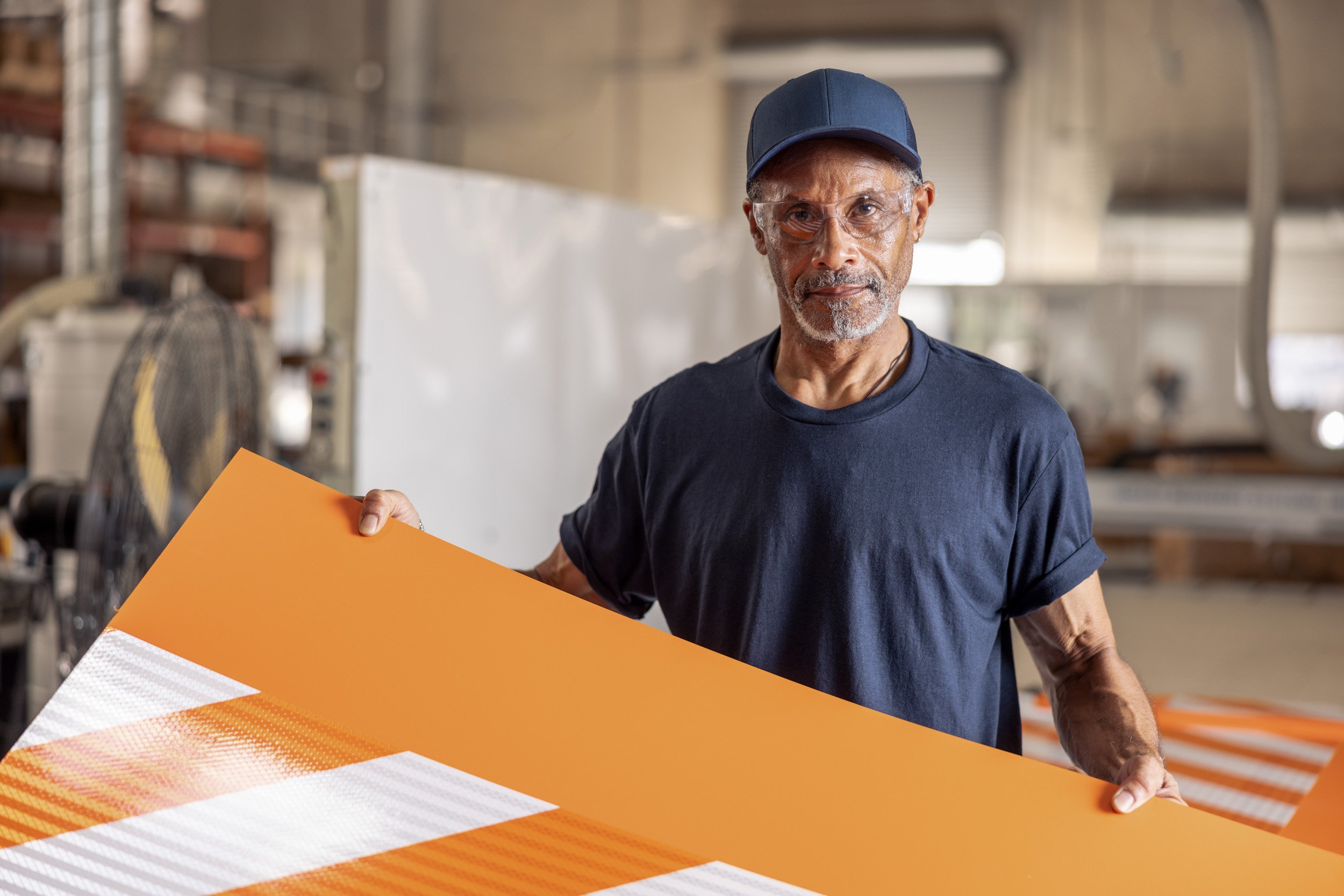 Older man wearing safety glasses and a blue cap holding a large orange and white striped sign in a factory or workshop setting.