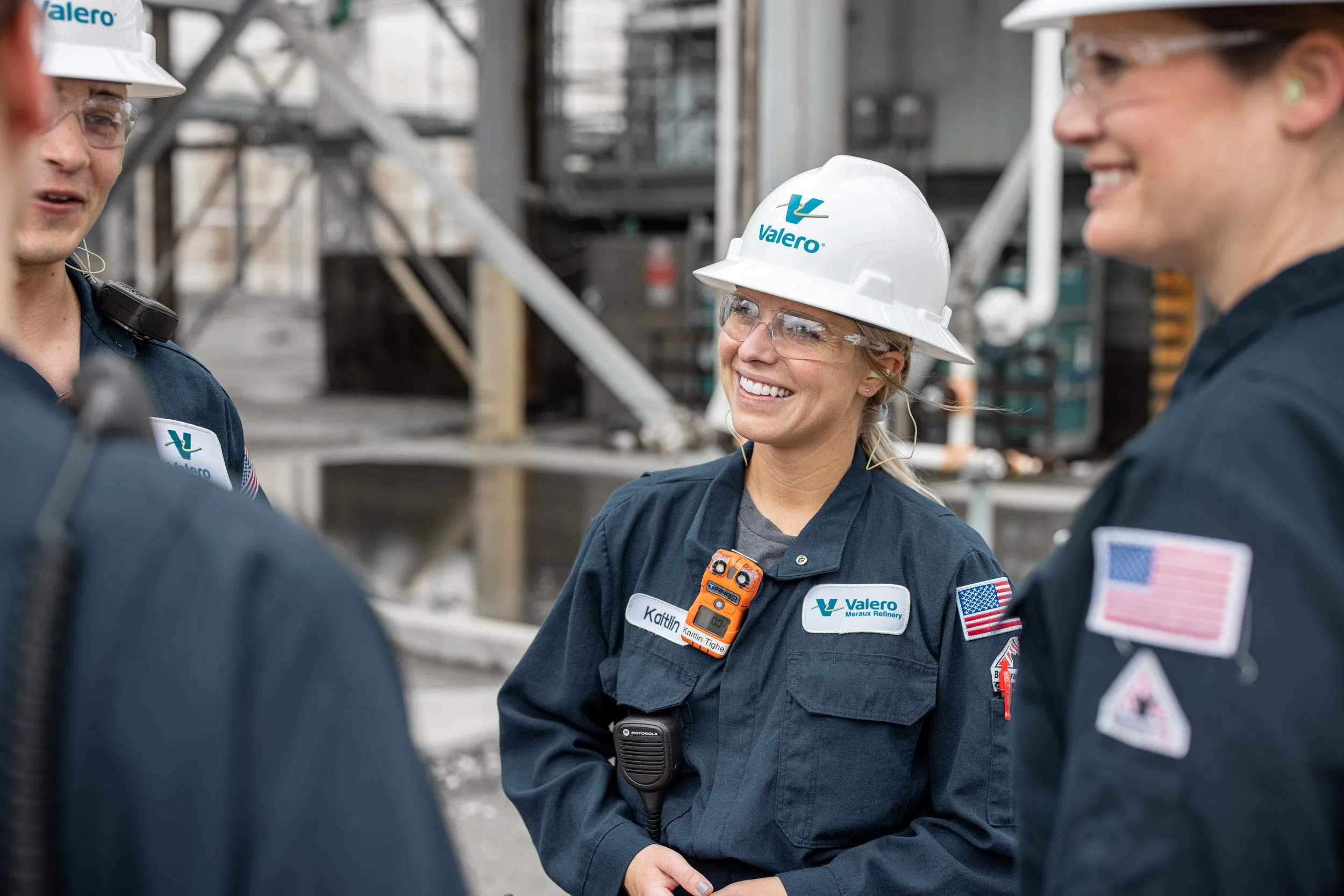 Three women in protective gear, including hard hats and safety glasses, standing in an industrial setting and smiling at each other. They are associated with Valero and Meraux Refinery, wearing uniforms with name tags and communication devices.