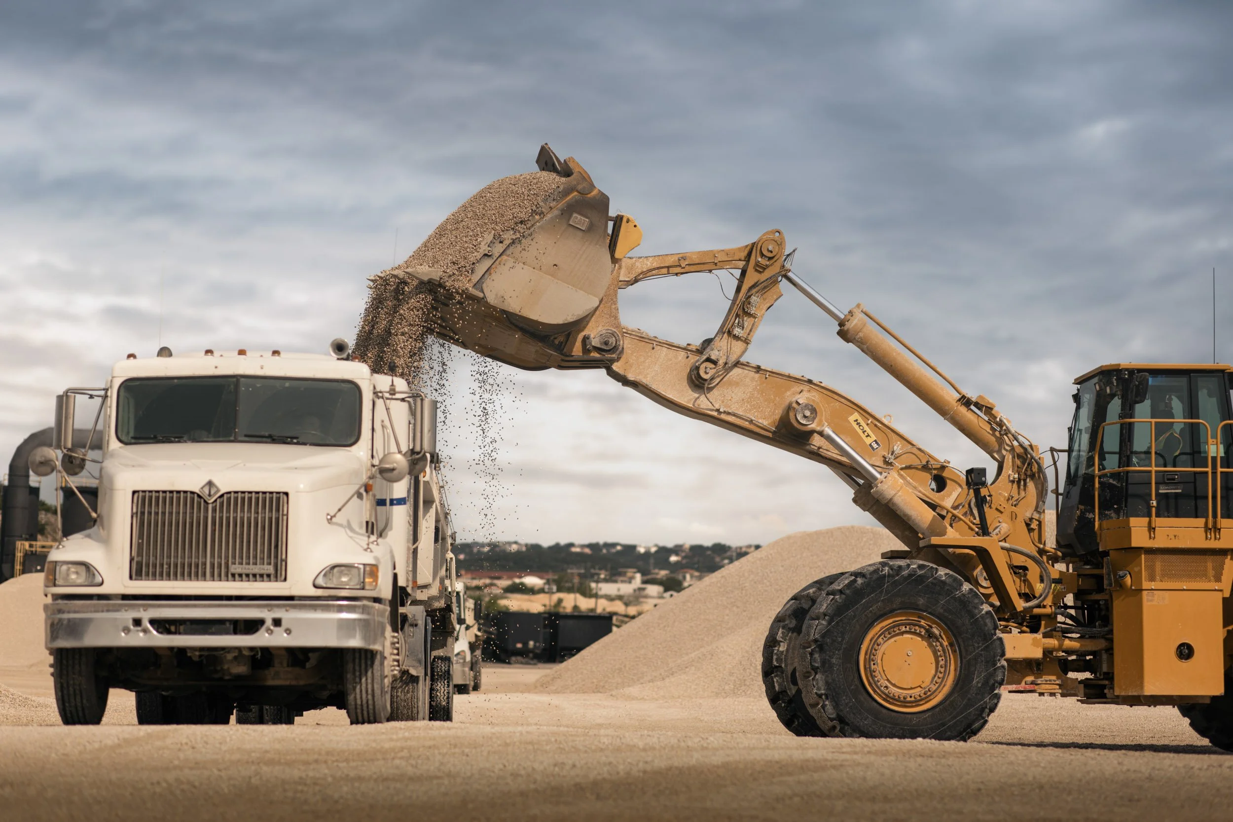 A construction site with a yellow excavator transferring gravel into a white dump truck under a cloudy sky.