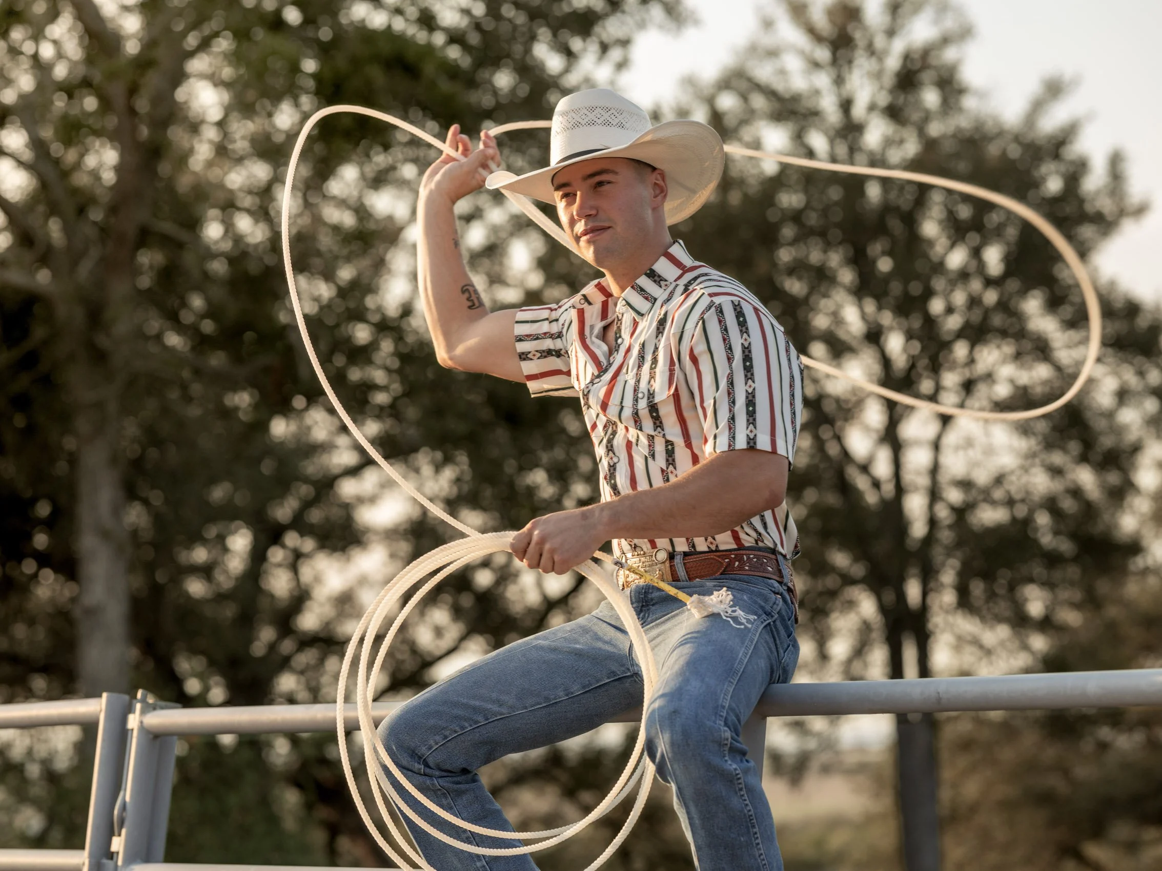 A man wearing a cowboy hat, striped shirt, and jeans, sitting on a metal railing outdoors, holding a lasso in one hand, with trees in the background.