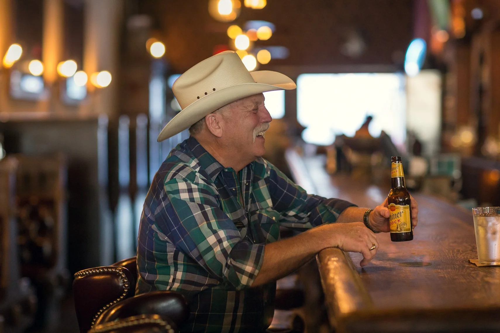 An older man with a cowboy hat and plaid shirt sitting at a bar, holding a beer bottle and laughing.