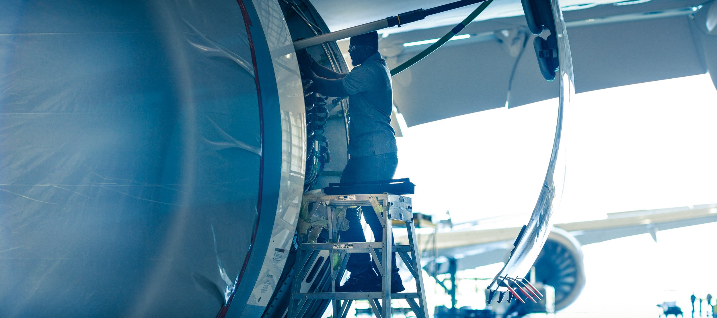 A person working on the cockpit of an aircraft with a ladder underneath, inside a hangar or large building.