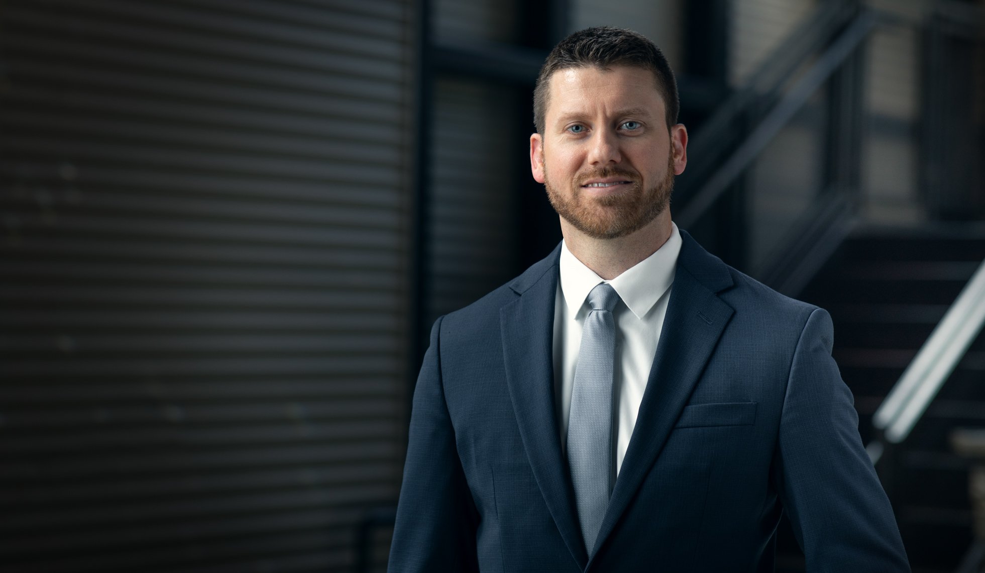 A man in a navy blue suit and grey tie standing outdoors with industrial buildings in the background.