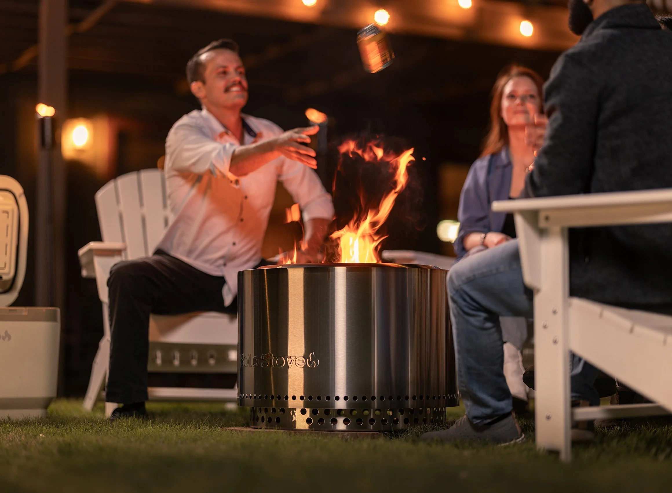 People sitting around a fire pit outdoors at night, with some throwing objects into the flames, in a social gathering.