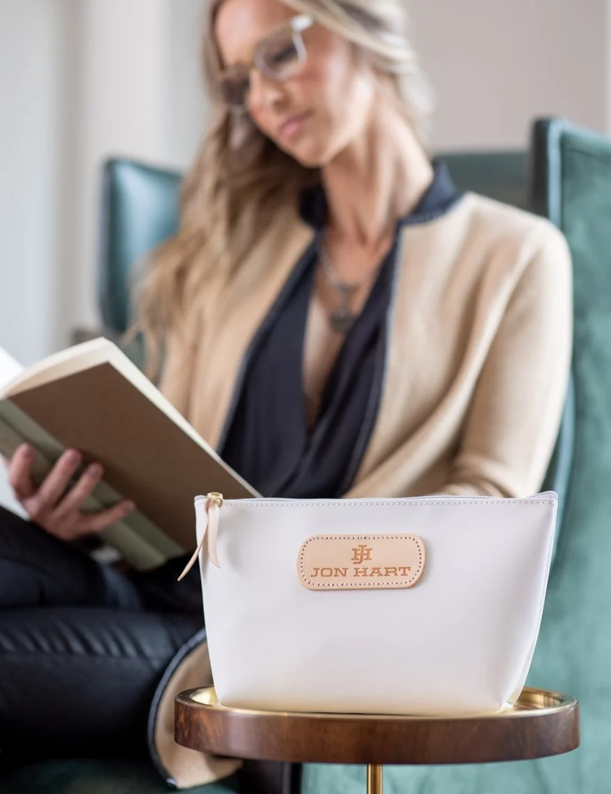 A woman with glasses reading a book, sitting on a couch with a Jon Hart cosmetic bag on a small round table in front of her.