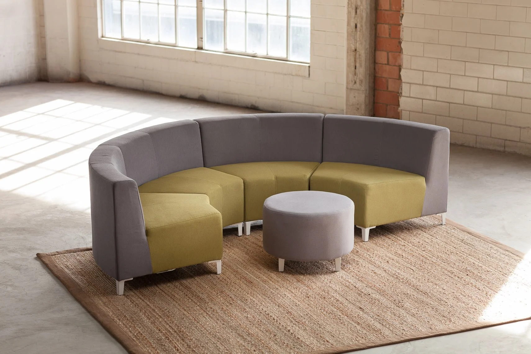 A modern gray and green sectional sofa with a round gray ottoman in a sunlit industrial-style room with exposed brick and white painted brick walls, and a large window, on a woven area rug.