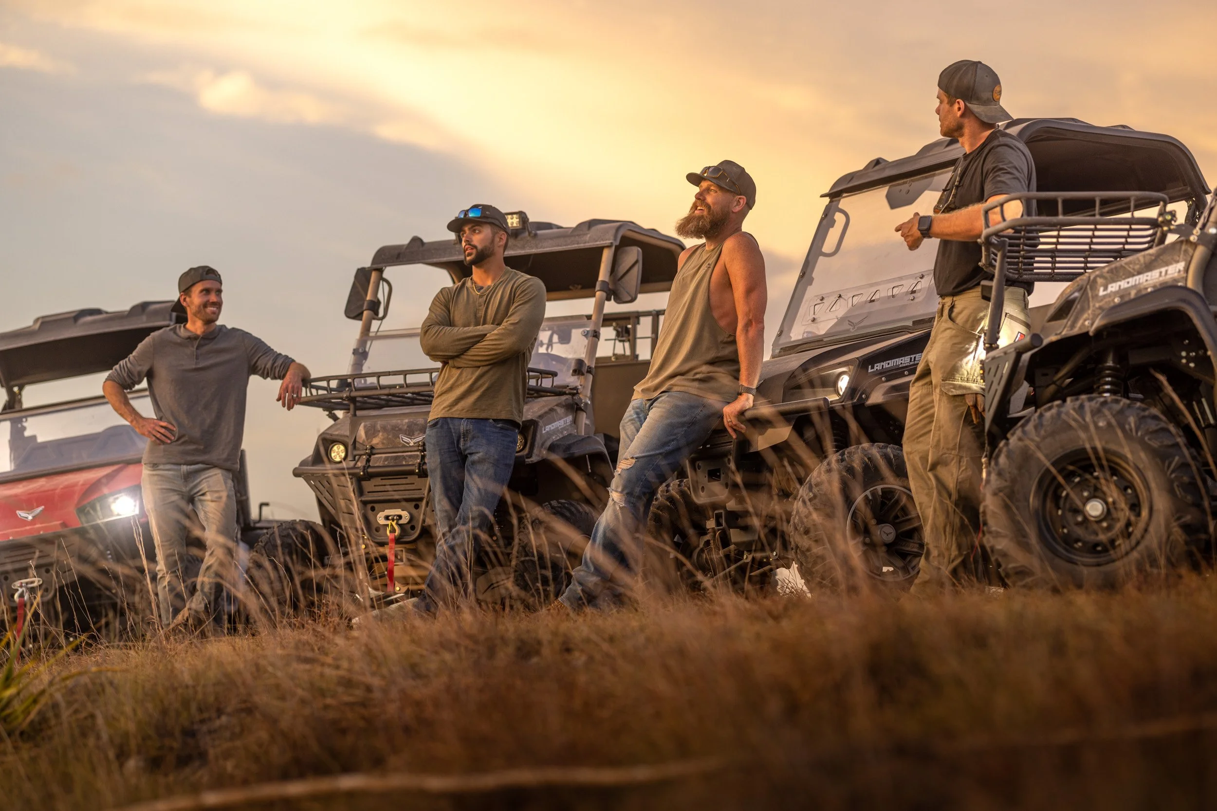 Four men stand near off-road utility vehicles in a field at sunset, engaging in conversation.