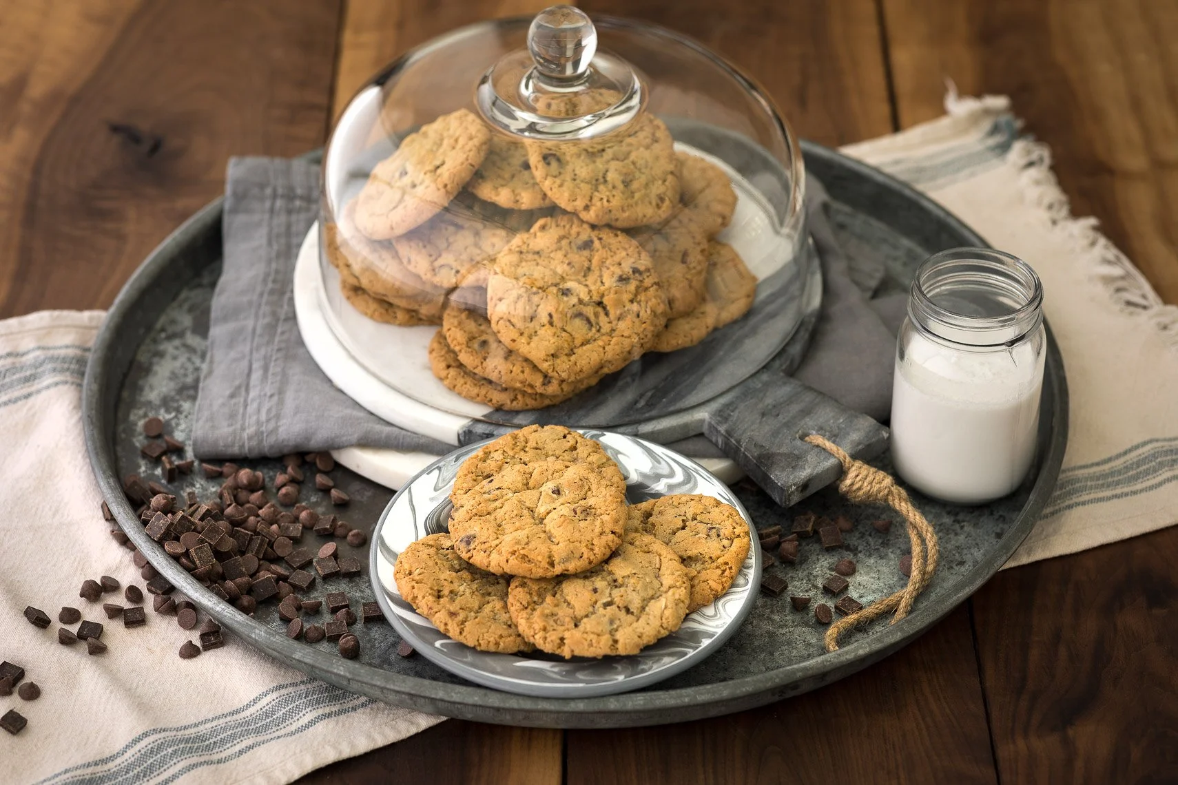 Chocolate chip cookies on a gray tray with a glass cover, a small pile of cookies on a decorative plate, chocolate chips scattered on a white cloth, and a jar of milk on a wooden table.