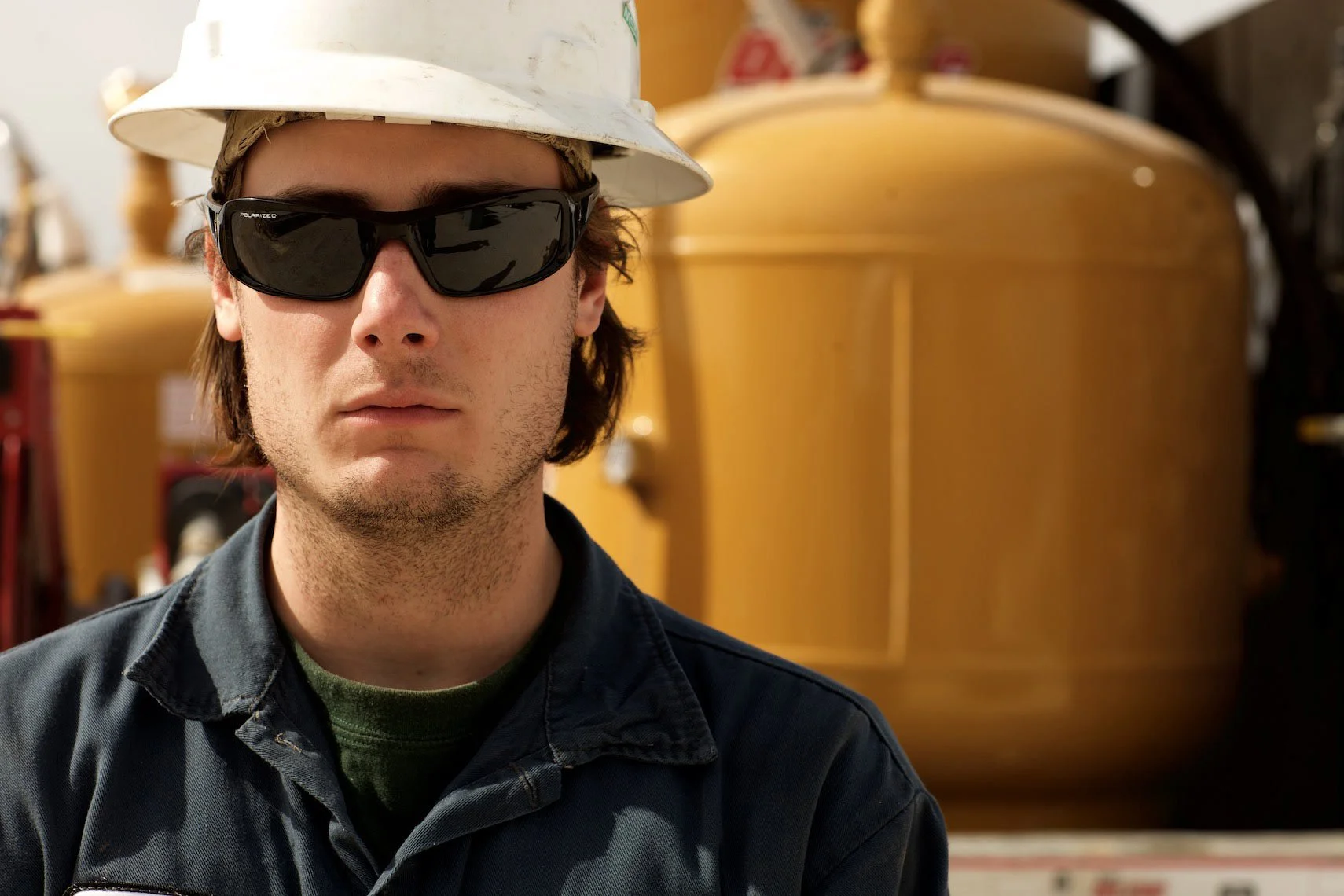 A man wearing safety glasses, a white hard hat, and a dark jacket, standing indoors in front of large yellow industrial tanks.