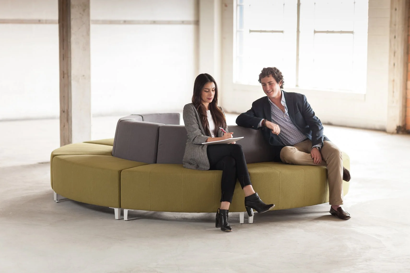 A man and woman sit on a modern yellow and gray couch in a spacious, unfinished room with large windows and exposed wooden beams. The woman is writing in a notebook, and the man is smiling and looking at her.