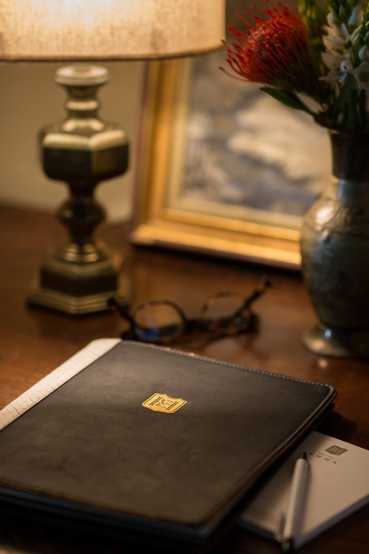 A desk with a closed black leather notebook, glasses, a pen, a small notepad, a lamp, a framed painting, and a vase with flowers.