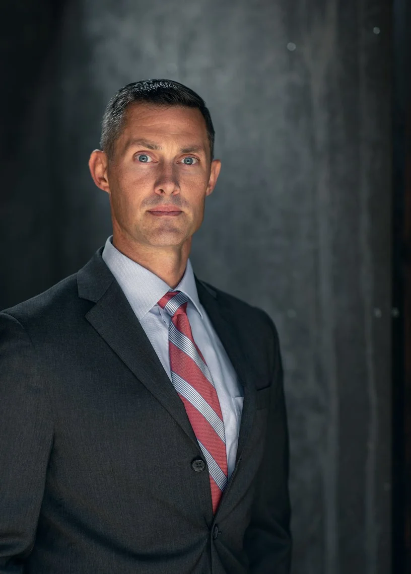 A serious-looking man with short dark hair, wearing a dark gray suit, light blue shirt, and red, white, and gray striped tie, standing against a dark background.