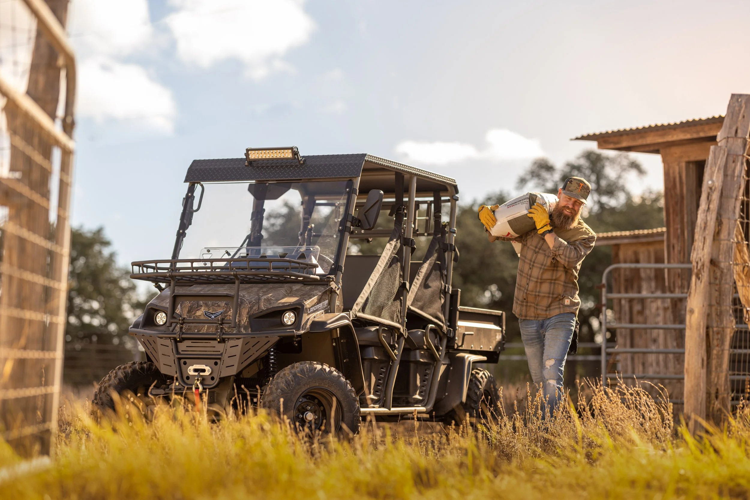 A man with a beard wearing a plaid shirt, jeans, and a cap, carrying a bag of cement next to a utility vehicle on a farm or construction site, with wooden structures nearby and a partly cloudy sky in the background.