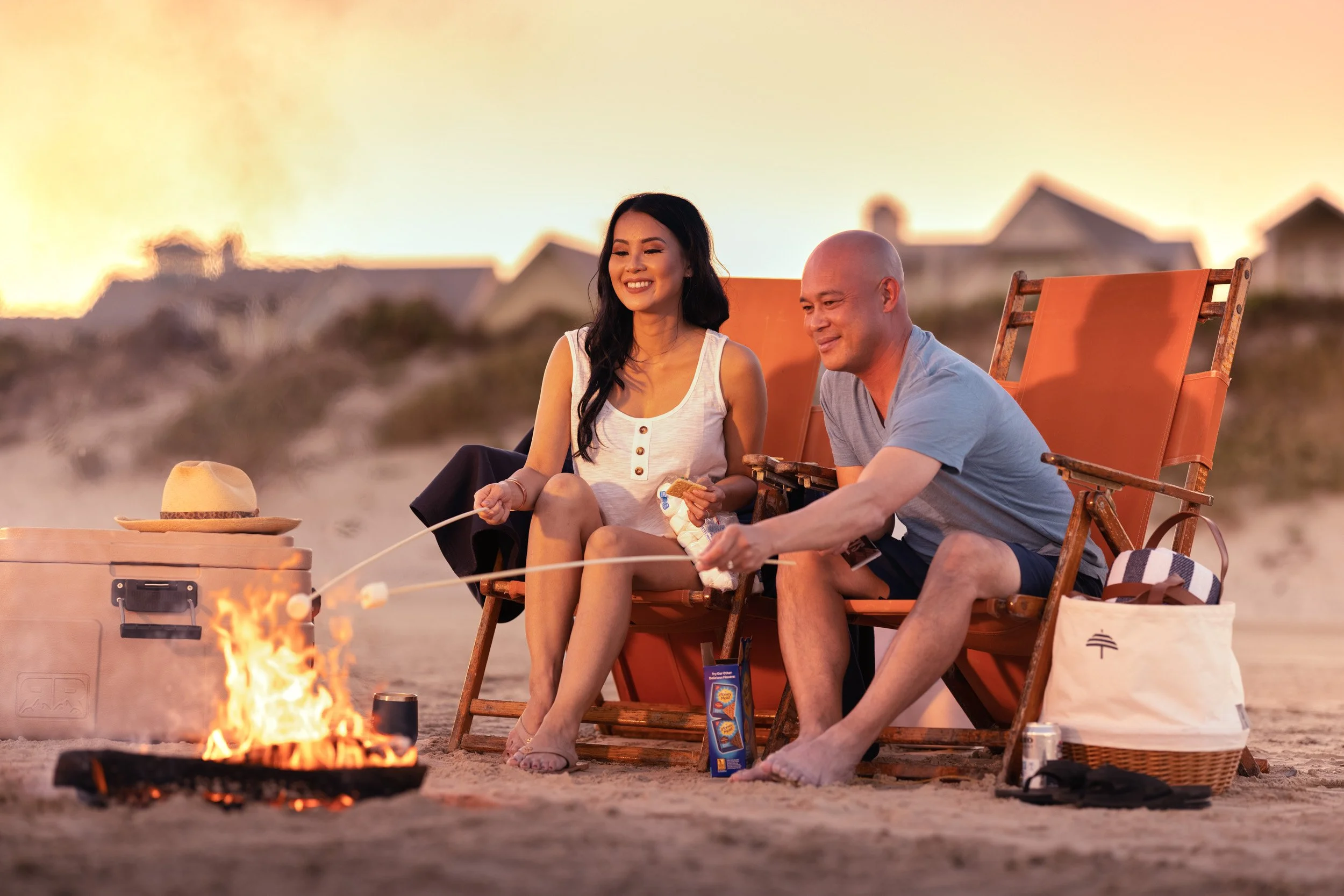 A woman and a man sitting on beach chairs by a campfire at sunset, roasting marshmallows.