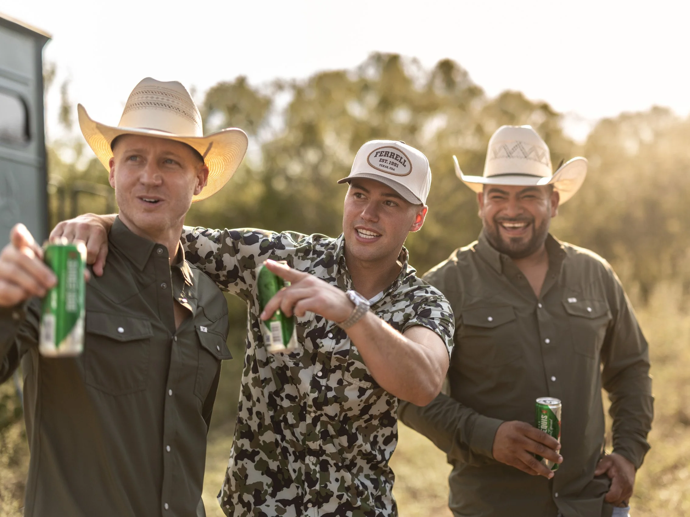Three men wearing cowboy hats and casual shirts, smiling and holding drinks outdoors during the daytime, with blurred green trees in the background.
