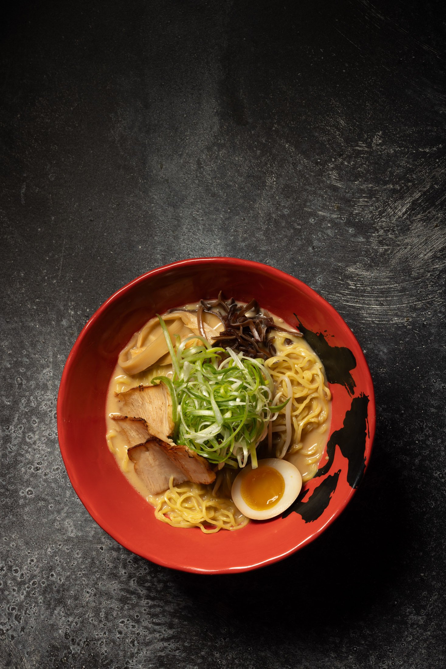 A bowl of ramen noodles topped with sliced pork, green onions, wood ear mushrooms, half an egg, and bamboo shoots, in a red bowl on a black textured surface.