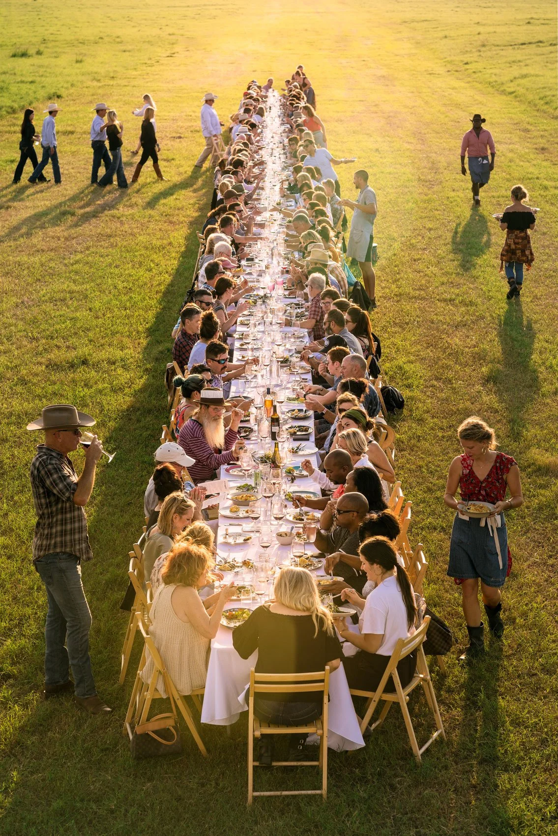 Long outdoor banquet table set on green grass with people dining, wearing casual and cowboy-style clothing, in a field during sunset, with servers walking nearby.