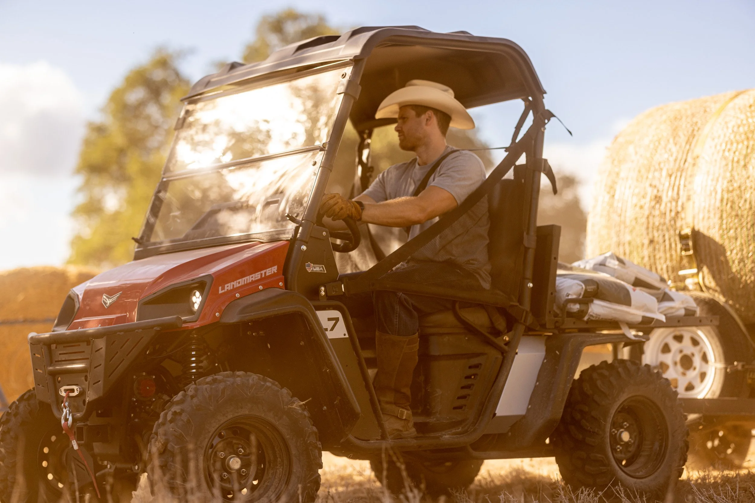 A man wearing a cowboy hat and gray t-shirt driving a red utility vehicle through a rural outdoor setting with hay bales and trees.