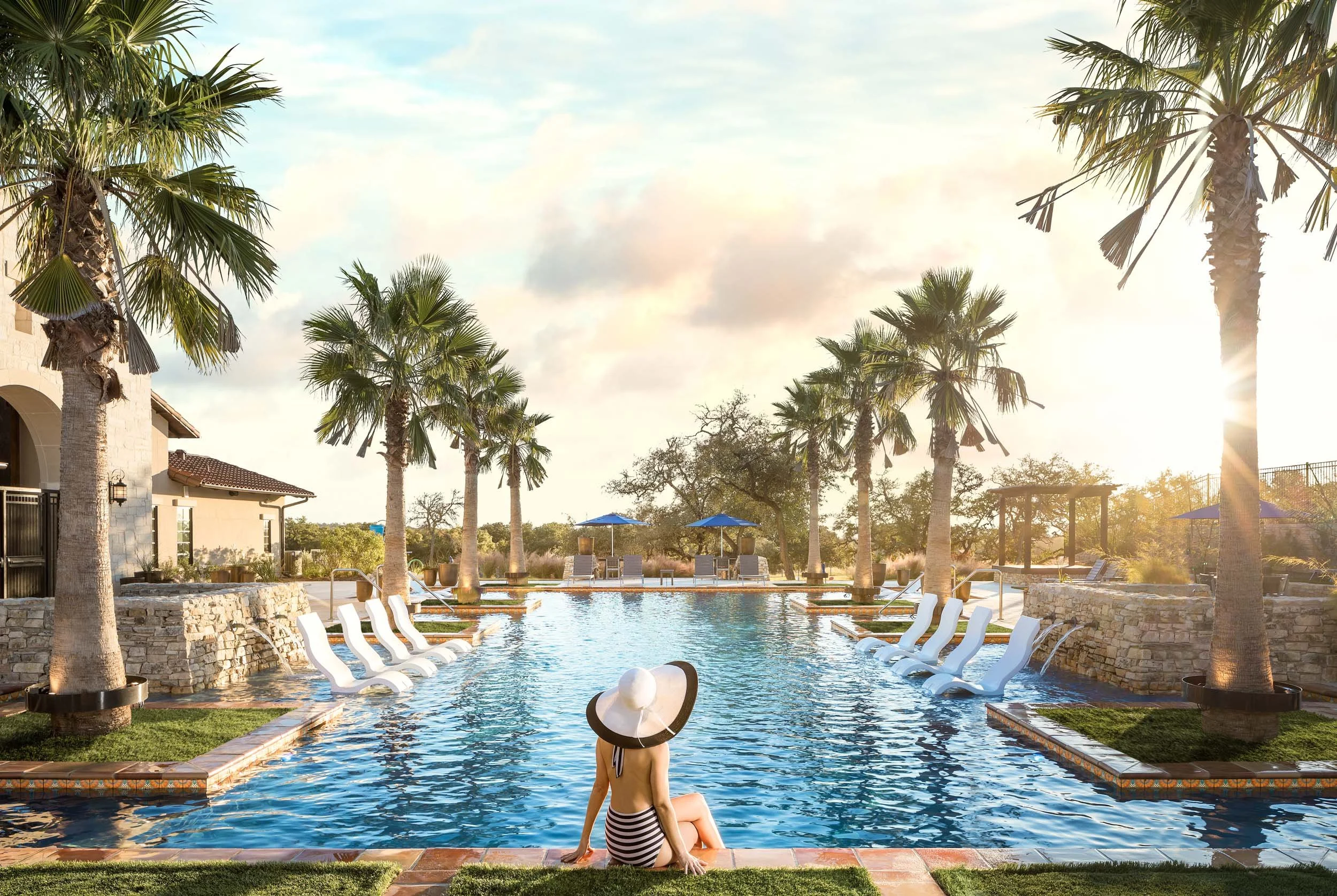 A woman sitting at the edge of a swimming pool with palm trees and a sky with clouds in the background, sun setting or rising casting a warm glow.