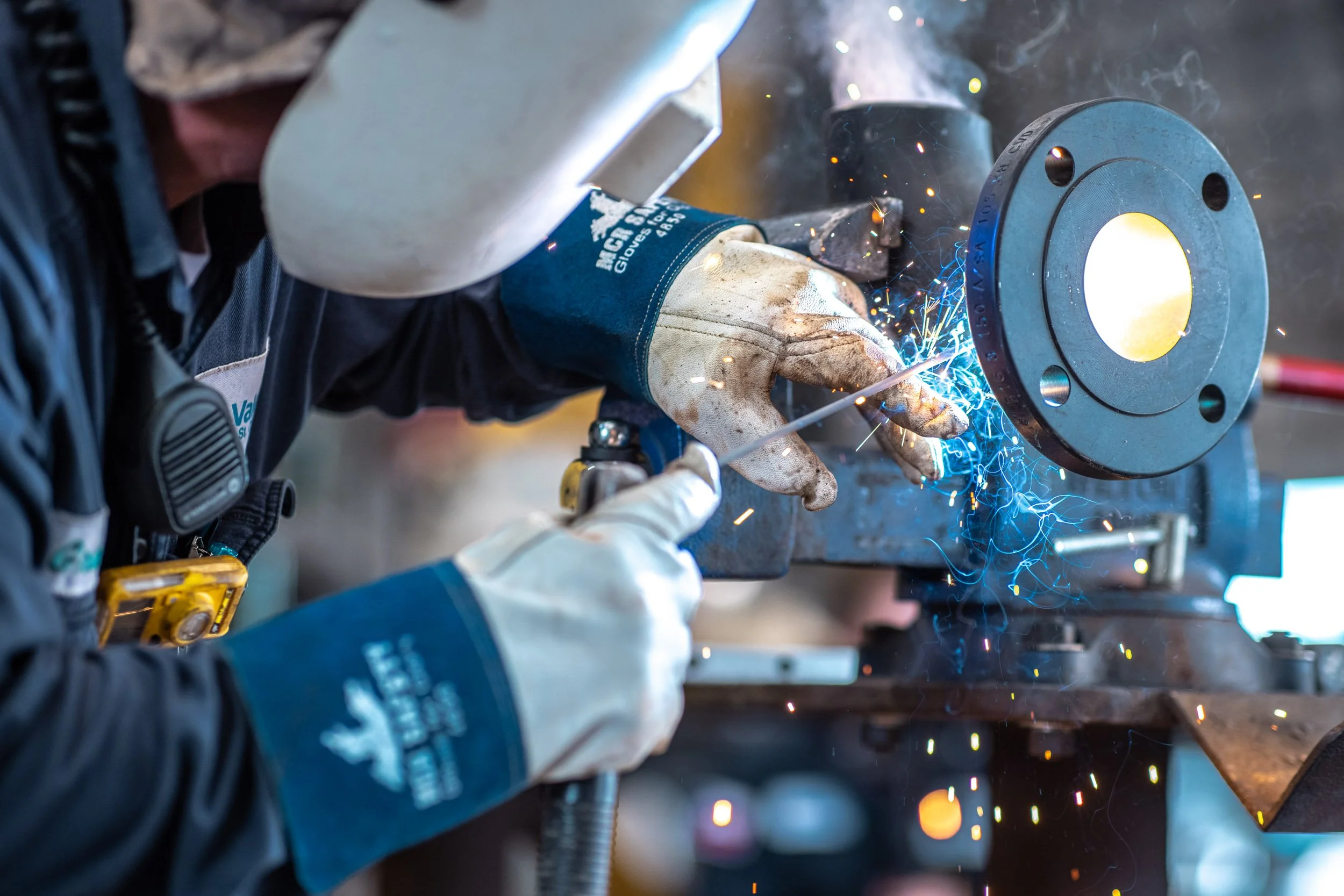 Worker welding metal pipe with sparks and bright light, wearing protective gloves and welding helmet.