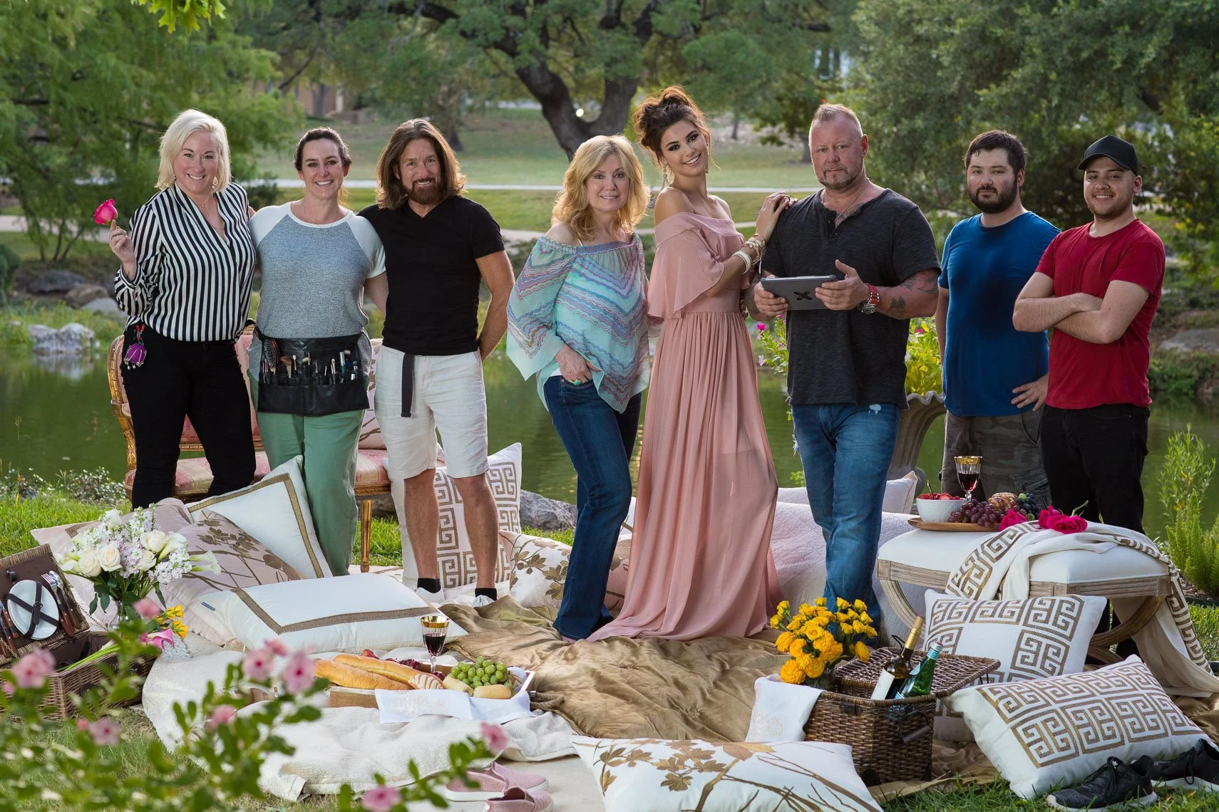 Group of eight people, six women and two men, standing outdoors near a lake or pond, surrounded by trees and greenery, with a picnic or outdoor gathering setup with cushions, pillows, flowers, and food in the foreground.