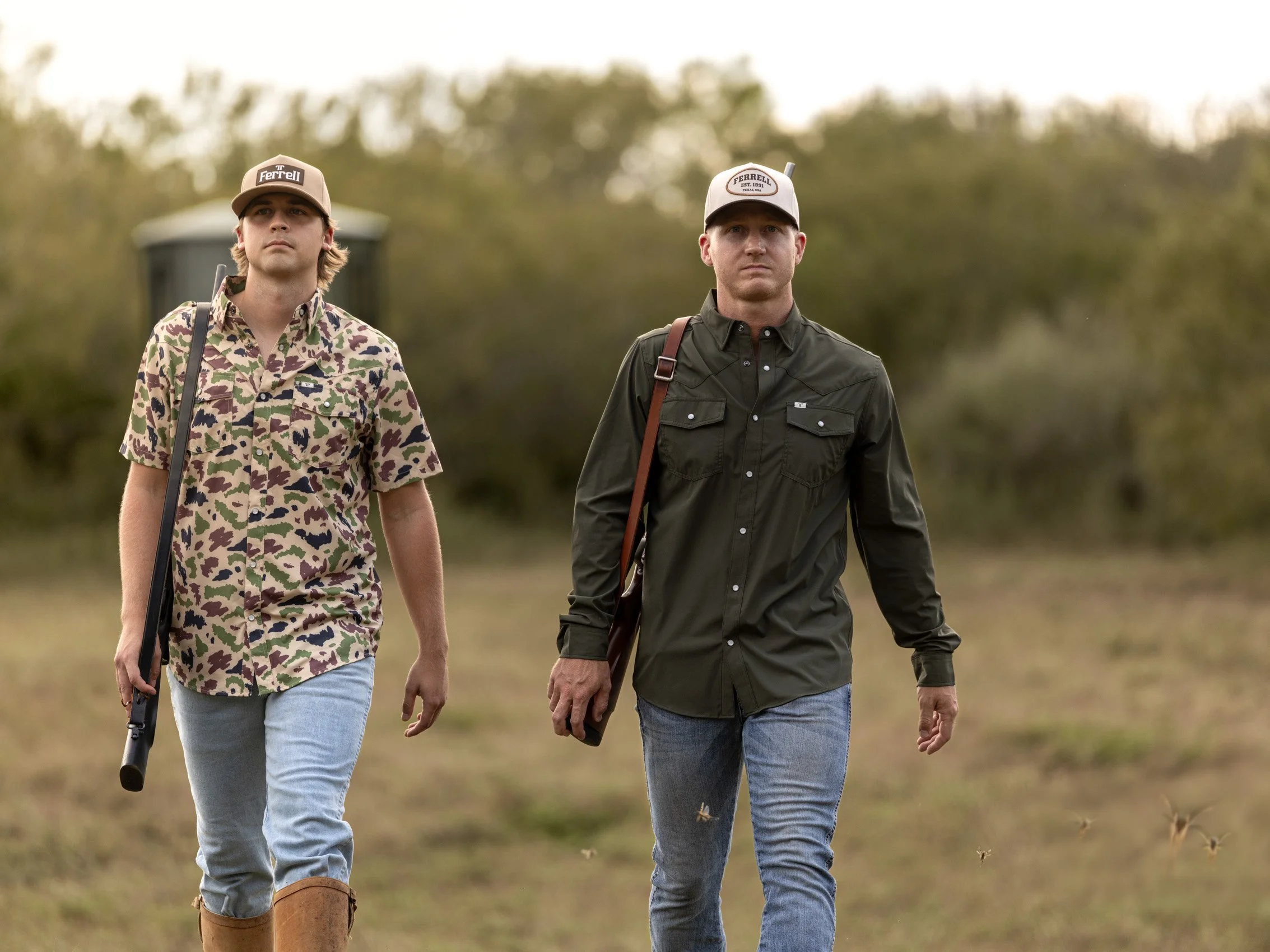 Two young men walking outdoors in a natural setting, dressed casually with one in a camo shirt and the other in a dark shirt, both wearing caps and carrying bags.
