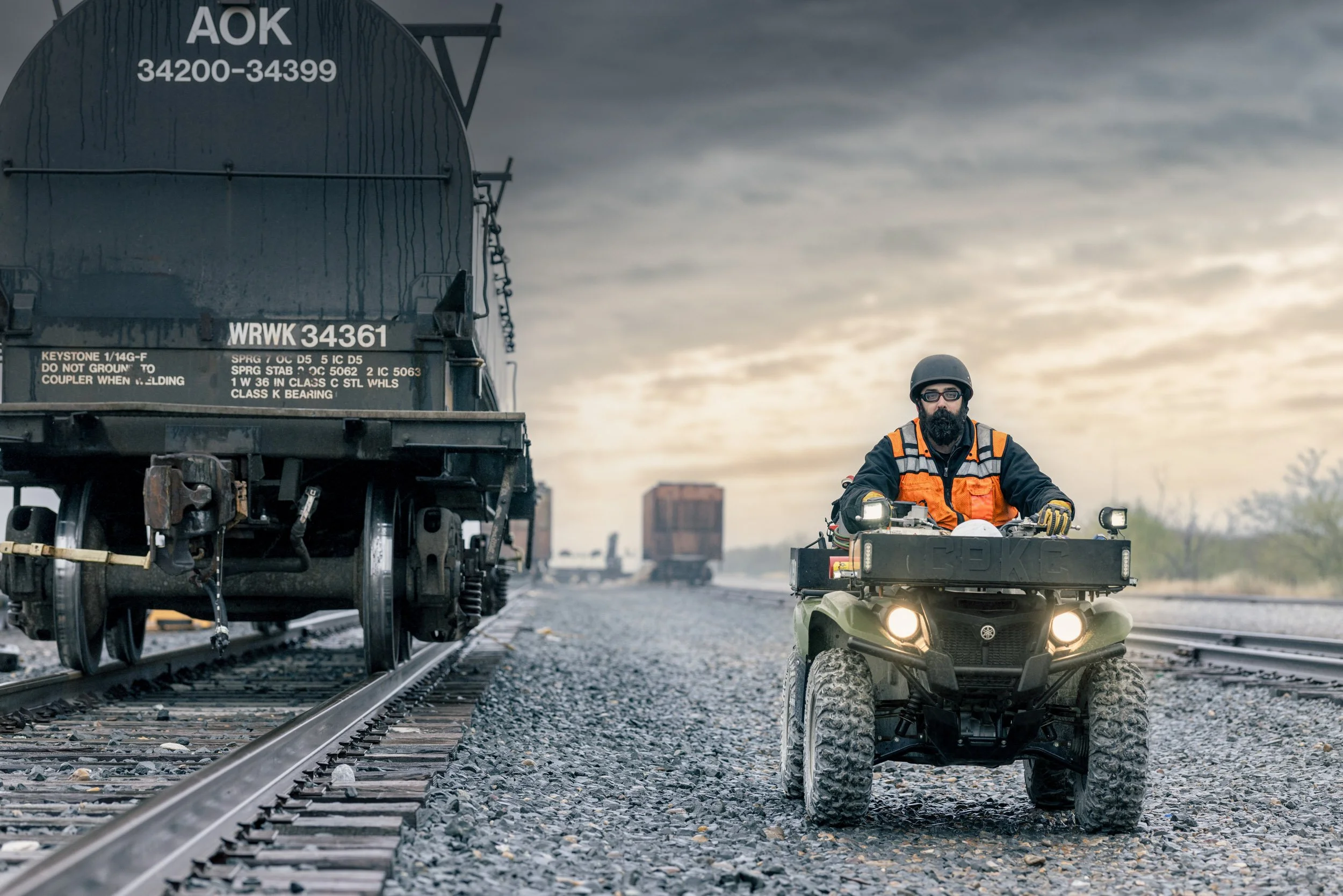 A man riding an ATV with a beard, glasses, and a black helmet, on train tracks during cloudy weather, with a large black railcar behind him and other trains in the background.