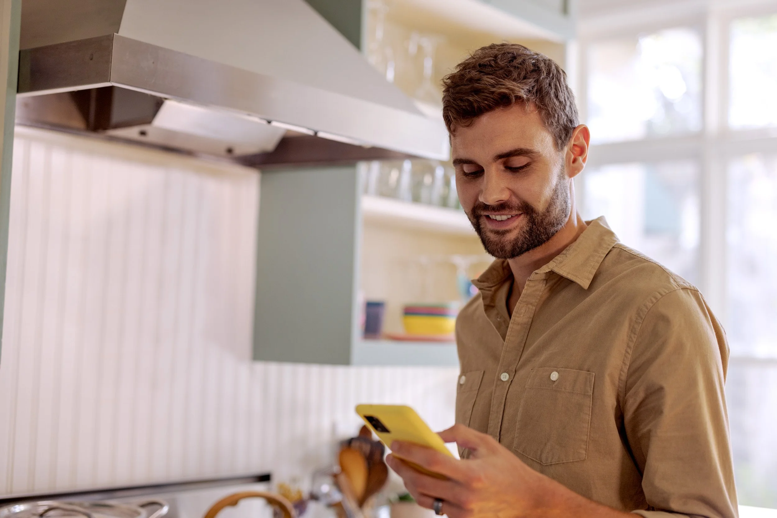 A man with short, curly brown hair and a beard, wearing a tan button-up shirt, is looking at his phone in a kitchen.