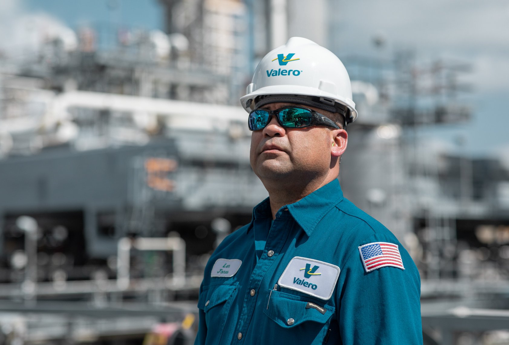 A man wearing a Valero uniform, white safety helmet, and reflective sunglasses standing in front of a marina with boats.