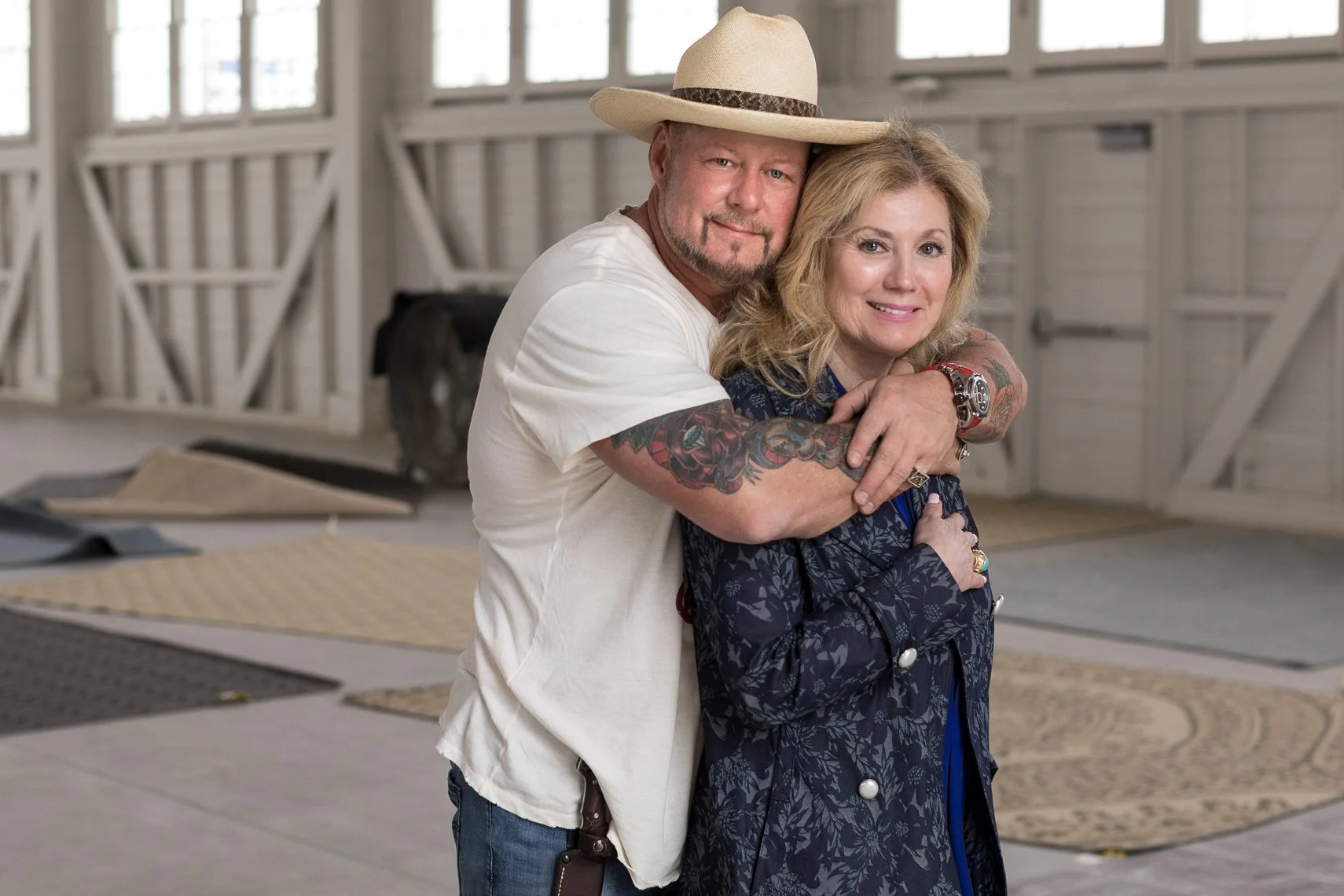 A man hugging a woman from behind inside a barn or garage with wooden walls and windows, both smiling at the camera.